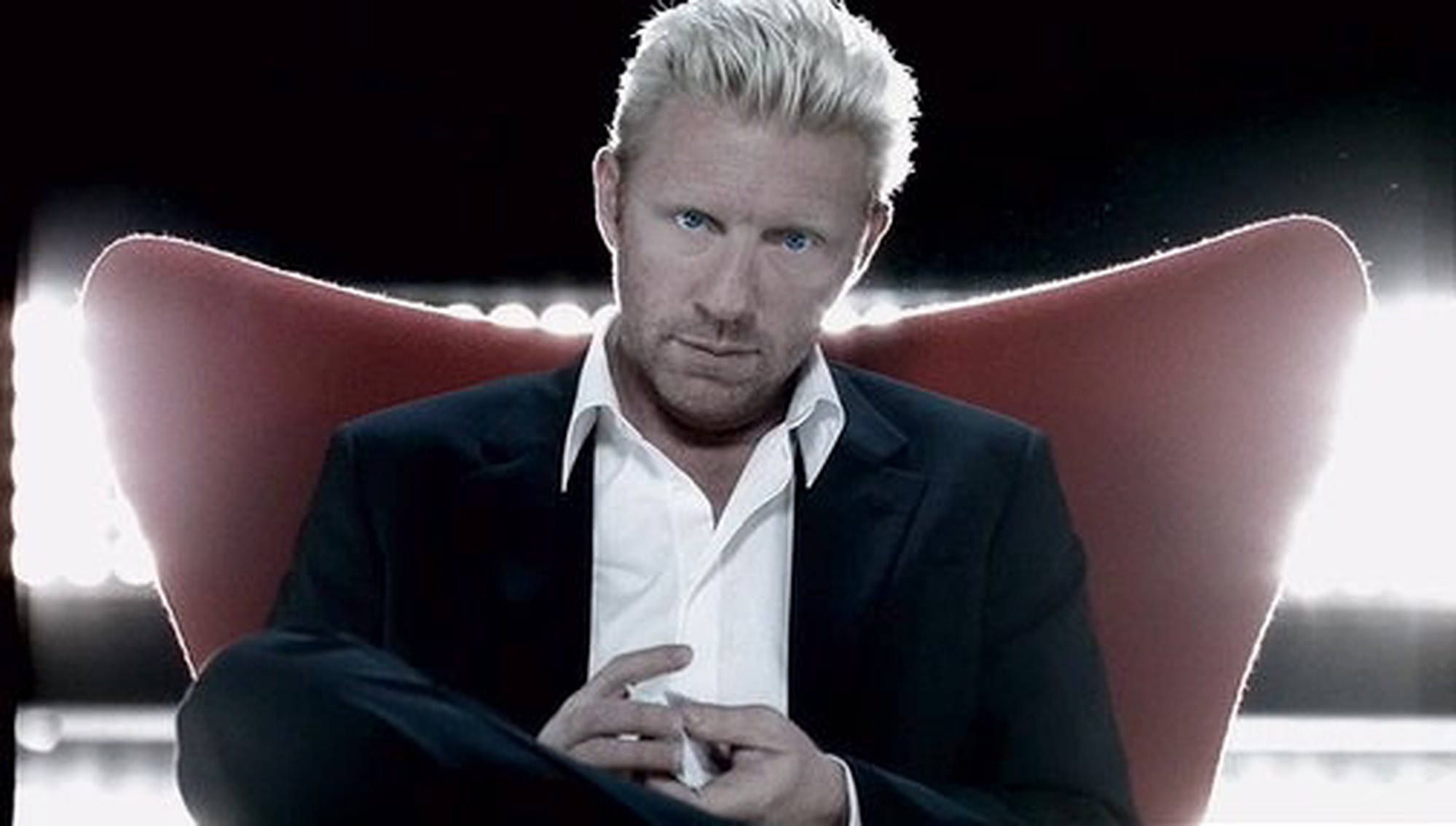 Boris Becker 桌布 壁紙 — 一名擁有鉑金色頭髮的男子坐在紅色椅子上，身穿黑色西裝和白色襯衫，背景昏暗，燈光明亮。