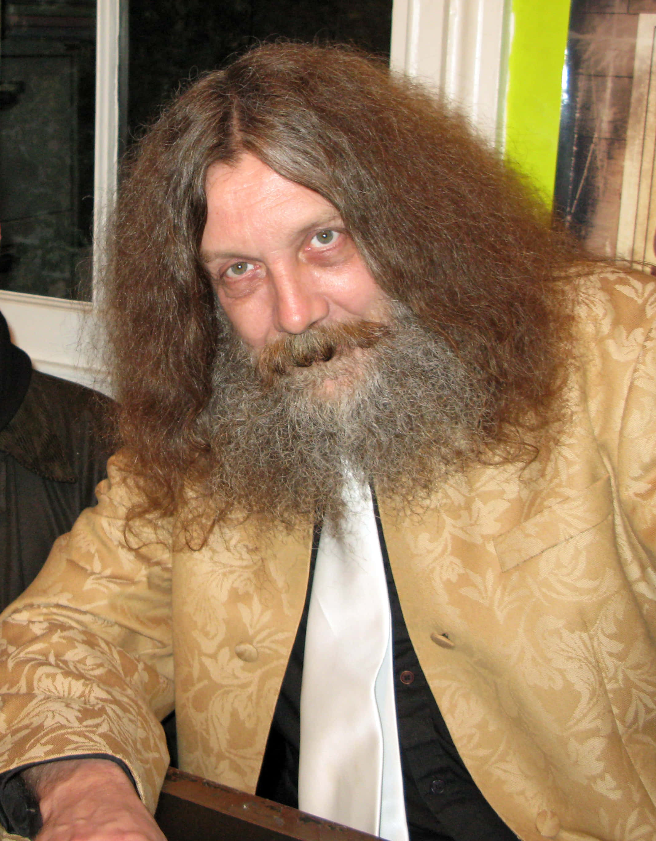 Alan Moore 桌布 壁紙 — 一名擁有長而波浪形棕色頭髮和濃密灰色鬍鬚的男子，穿著帶圖案的金色外套和白色領帶，正看著鏡頭。