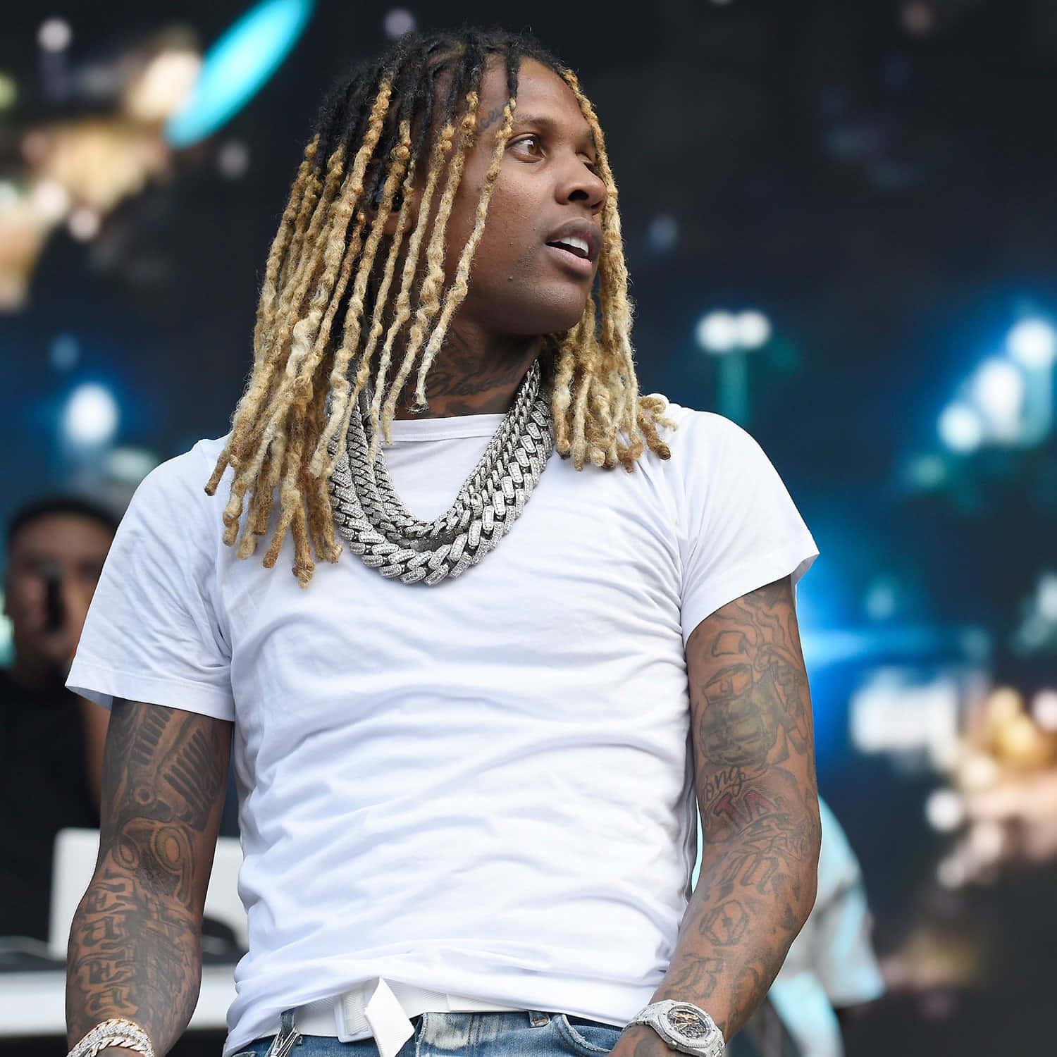Lil Durk 桌布 壁紙 — 一名擁有長金髮辮和沉重銀鏈的男子，穿著白色T恤和牛仔褲，在音樂會上望向鏡頭外。