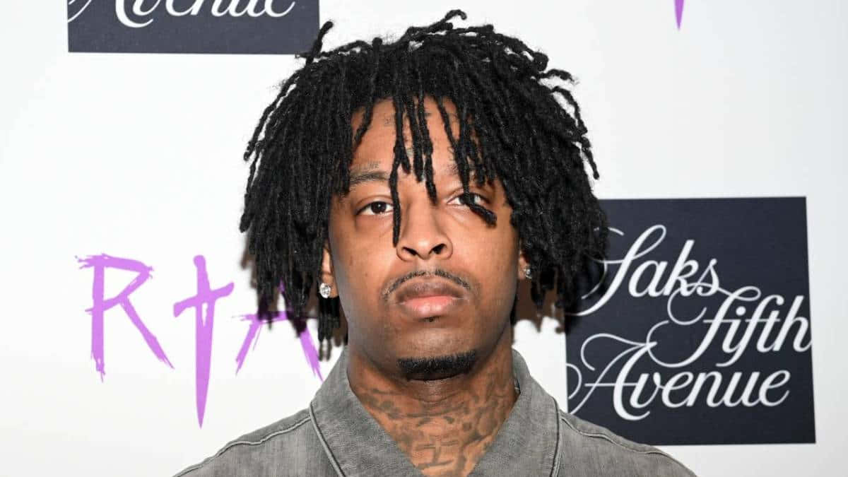 21 Savage 桌布 壁紙 — 一名擁有黑色辮子頭和可見頸部刺青的男子，在品牌活動背景前直視鏡頭。