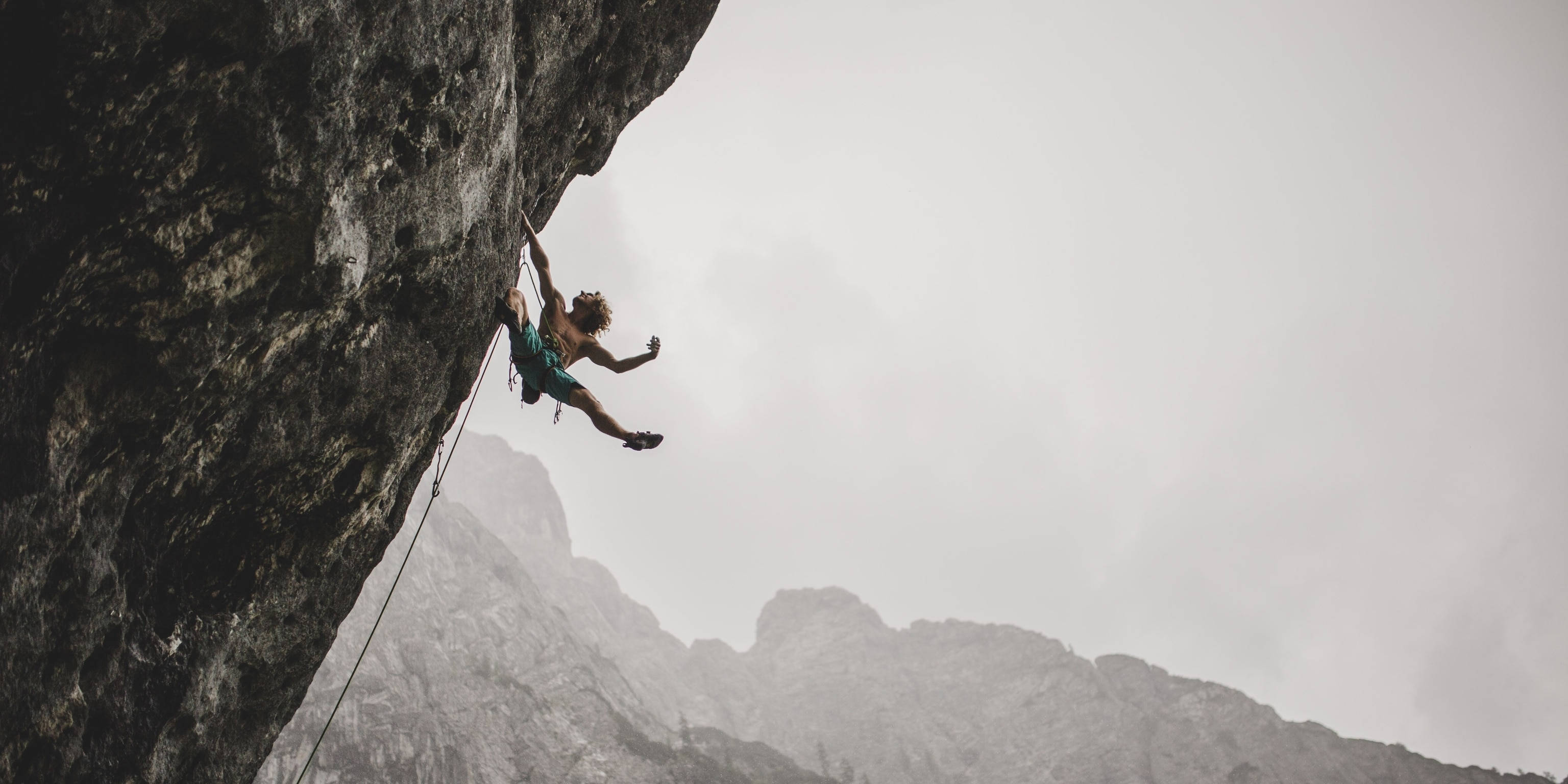 Climbing 桌布 壁紙 — 一名攀岩者在多雲的天空下，以霧氣瀰漫的山脈為背景，攀爬陡峭的岩壁。