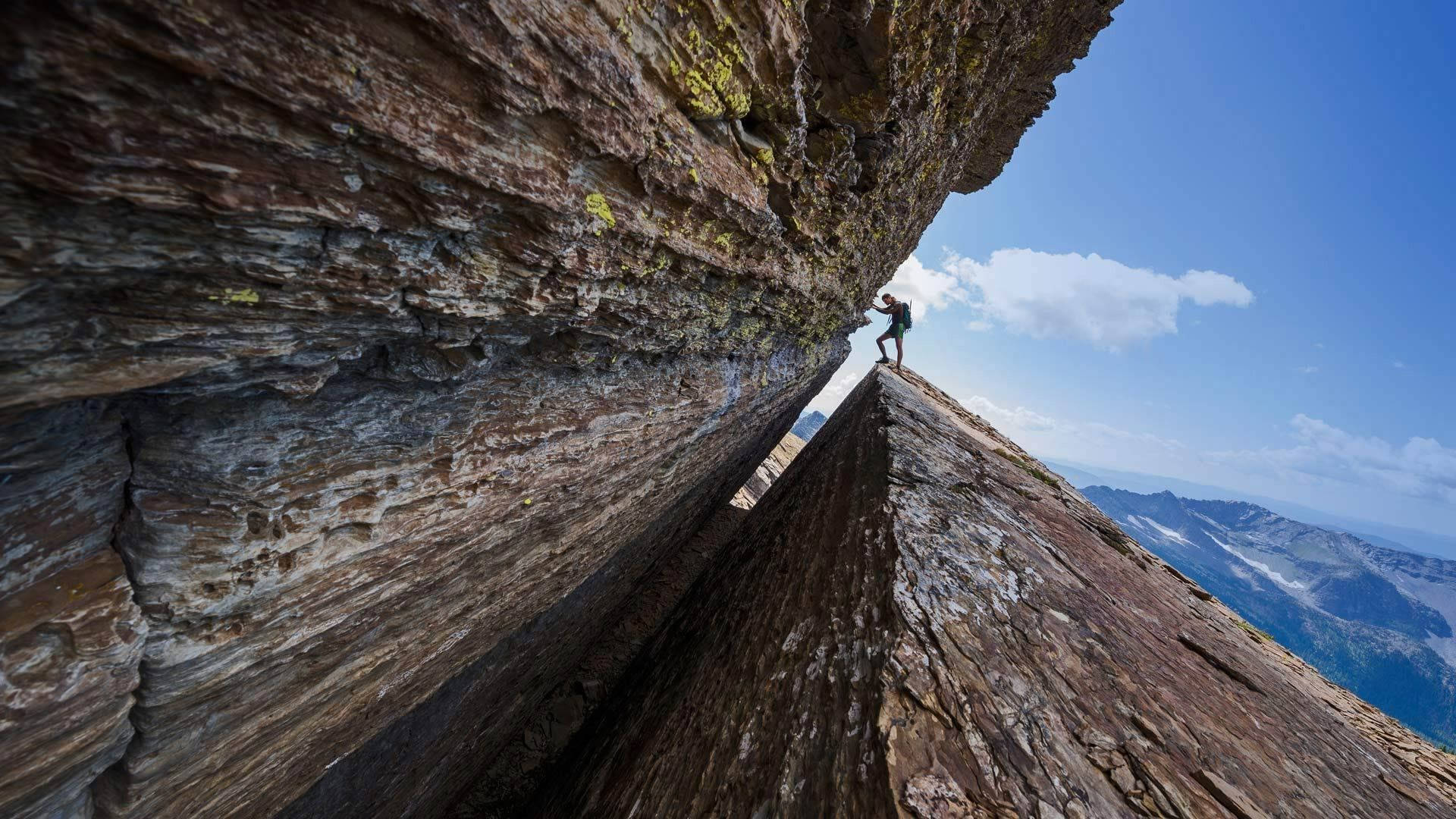 Climbing 桌布 壁紙 — 一名攀岩者在廣闊的藍天下攀登狹窄的岩石山脊，下方是遠處積雪的山峰。