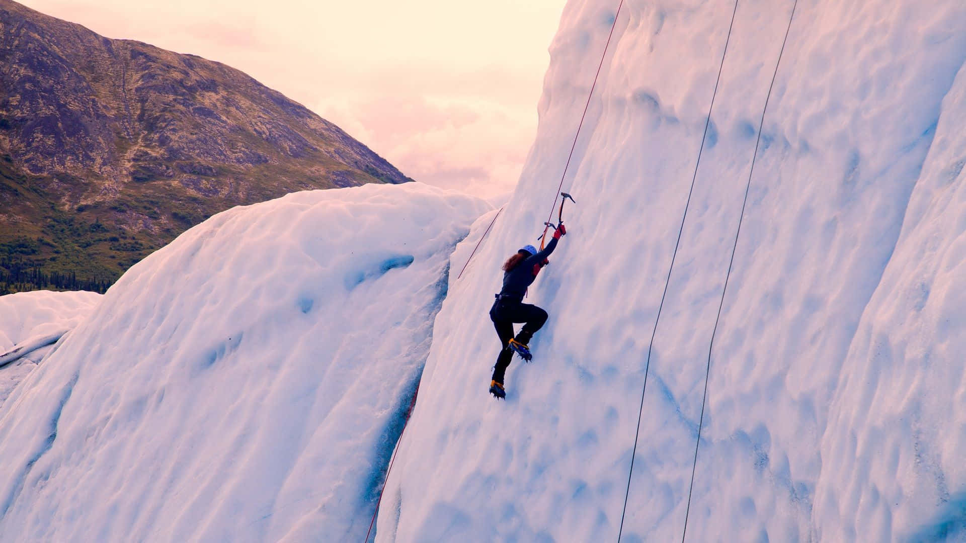 Ice Climbing 桌布 壁紙 — 一名攀岩者在日落時分用繩索和冰斧攀登高聳的冰川壁，背景是山巒。