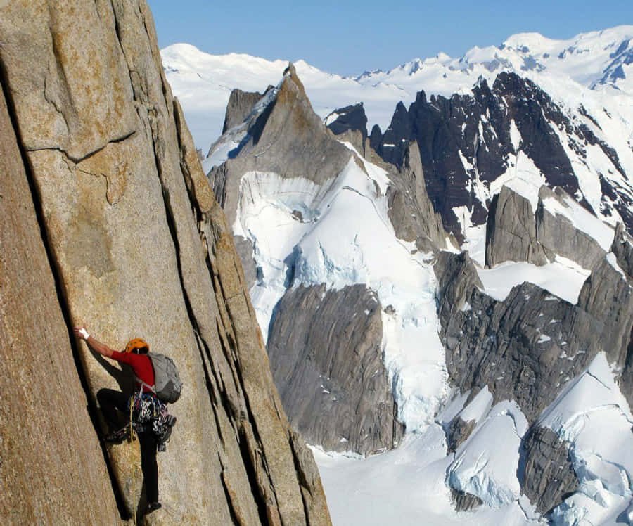 Patagonia 桌布 壁紙 — 一名攀岩者在晴朗的藍天下，以積雪覆蓋的鋸齒狀山峰為背景，攀爬陽光照耀的岩壁。