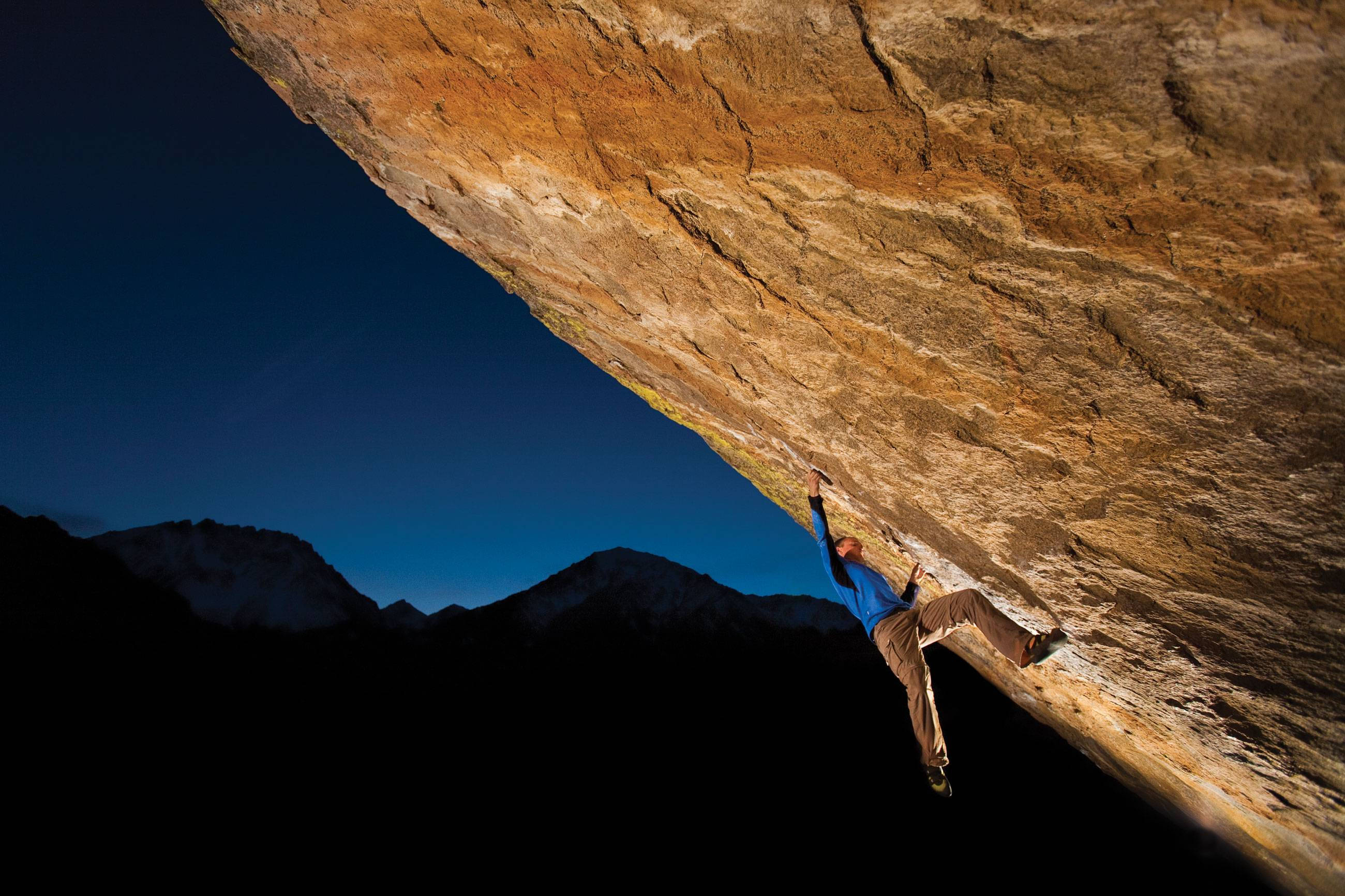 Climbing 桌布 壁紙 — 一名攀岩者在深藍色暮色天空下攀爬陡峭的懸垂岩壁，遠處白雪覆蓋的山峰在下方形成剪影。
