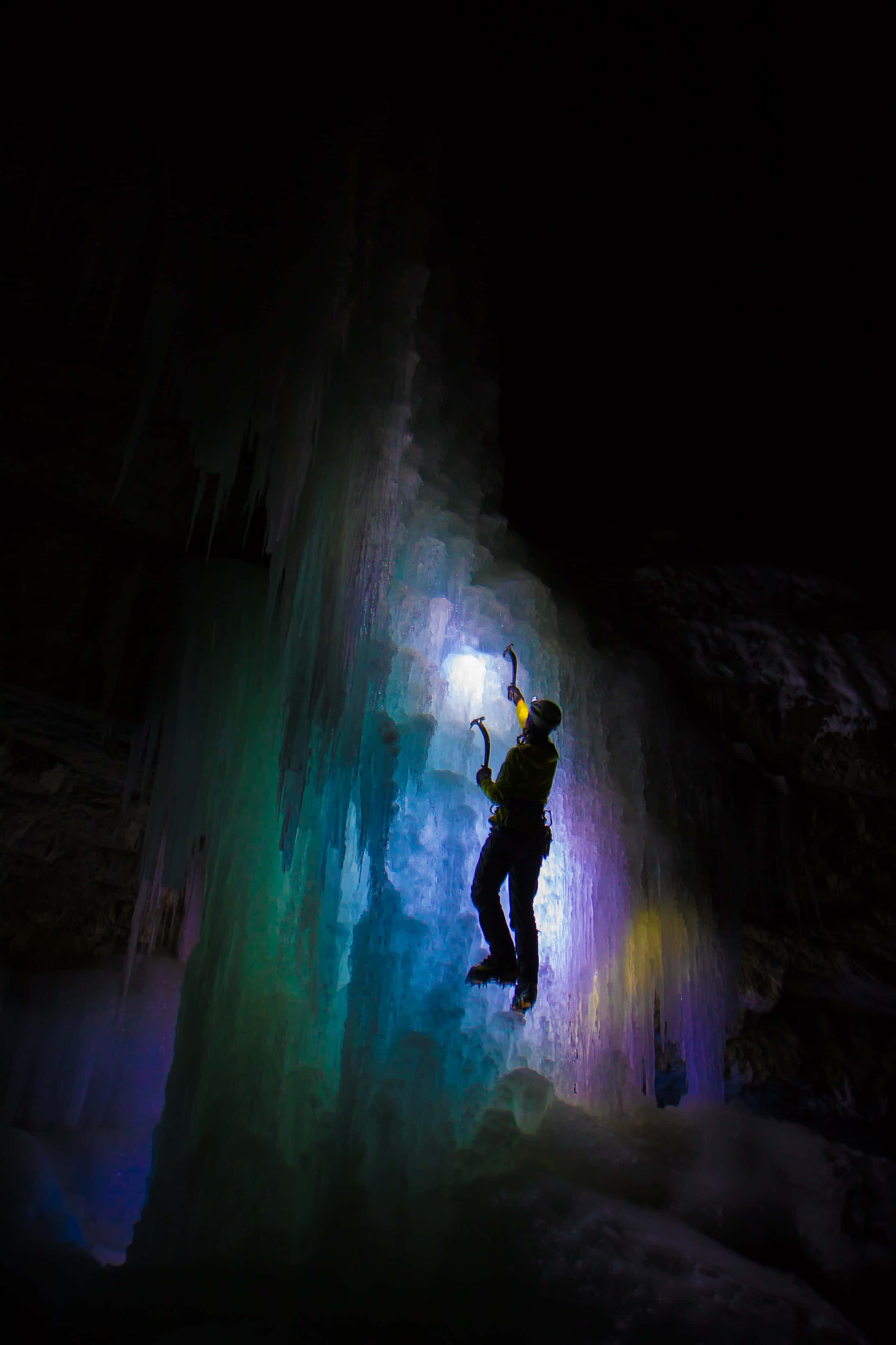 Ice Climbing 桌布 壁紙 — 一名攀岩者在黑暗洞穴中攀登由彩色燈光照亮的發光冰壁。