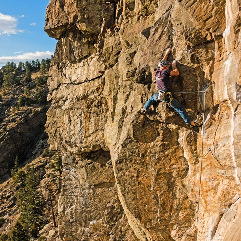 Climbing 桌布 壁紙 — 一名攀岩者帶著裝備和繩索，在松樹和藍天的背景下，攀登崎嶇的陽光岩壁。