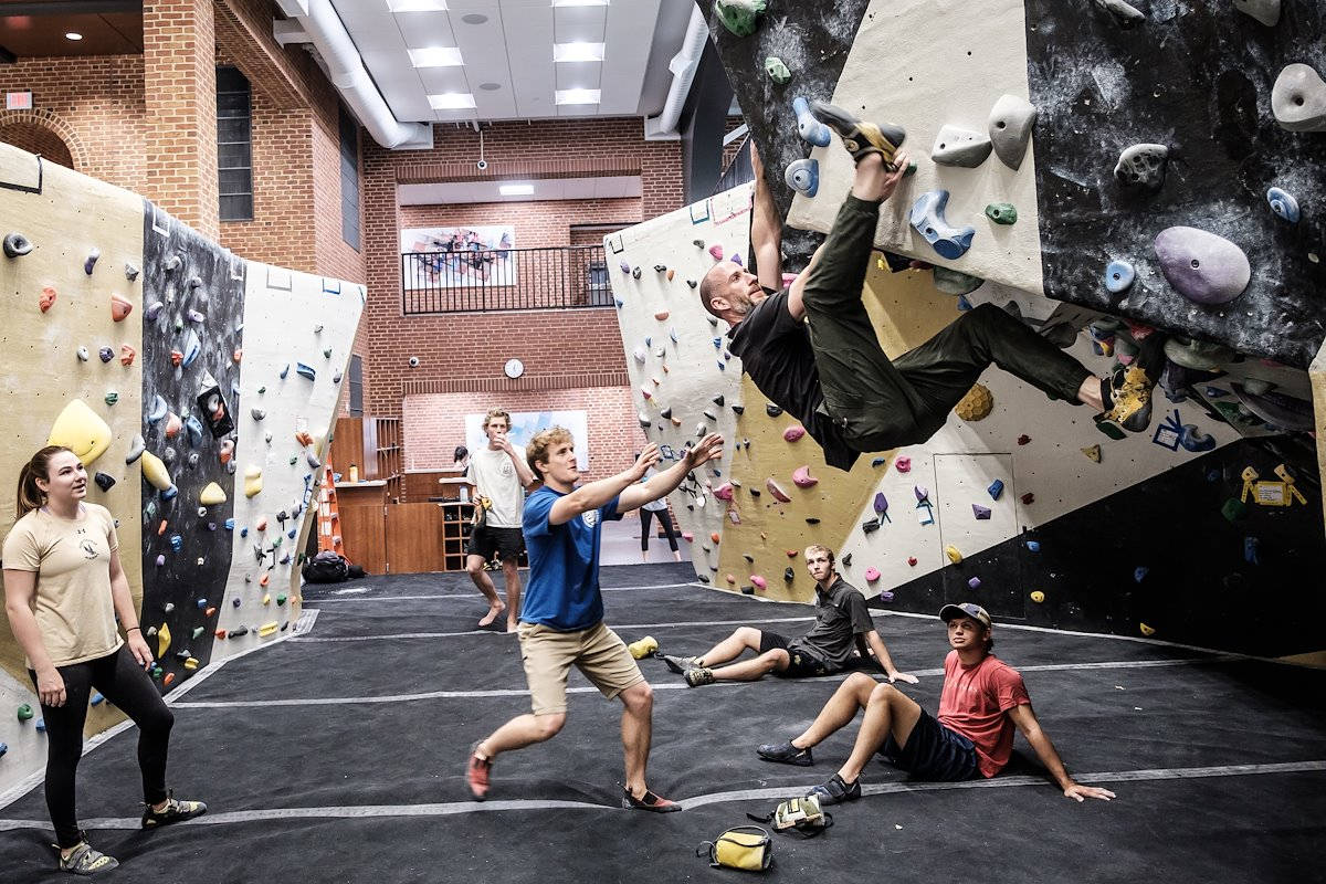 Climbing 桌布 壁紙 — 一名攀岩者懸掛在室內攀岩牆的半空中，教練做出鼓勵的手勢，而其他人在下方觀看或休息。