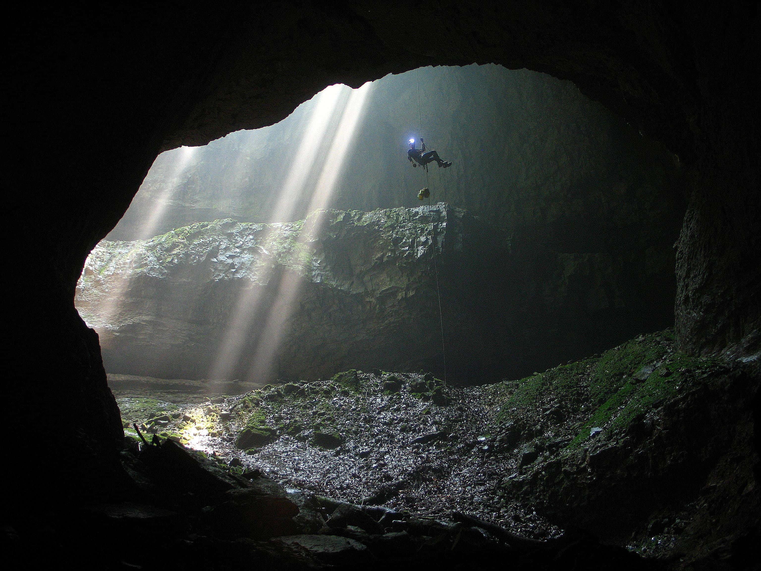 Cave 桌布 壁紙 — 一名攀岩者懸掛在廣闊的黑暗洞穴中，陽光從上方的開口穿透而入。
