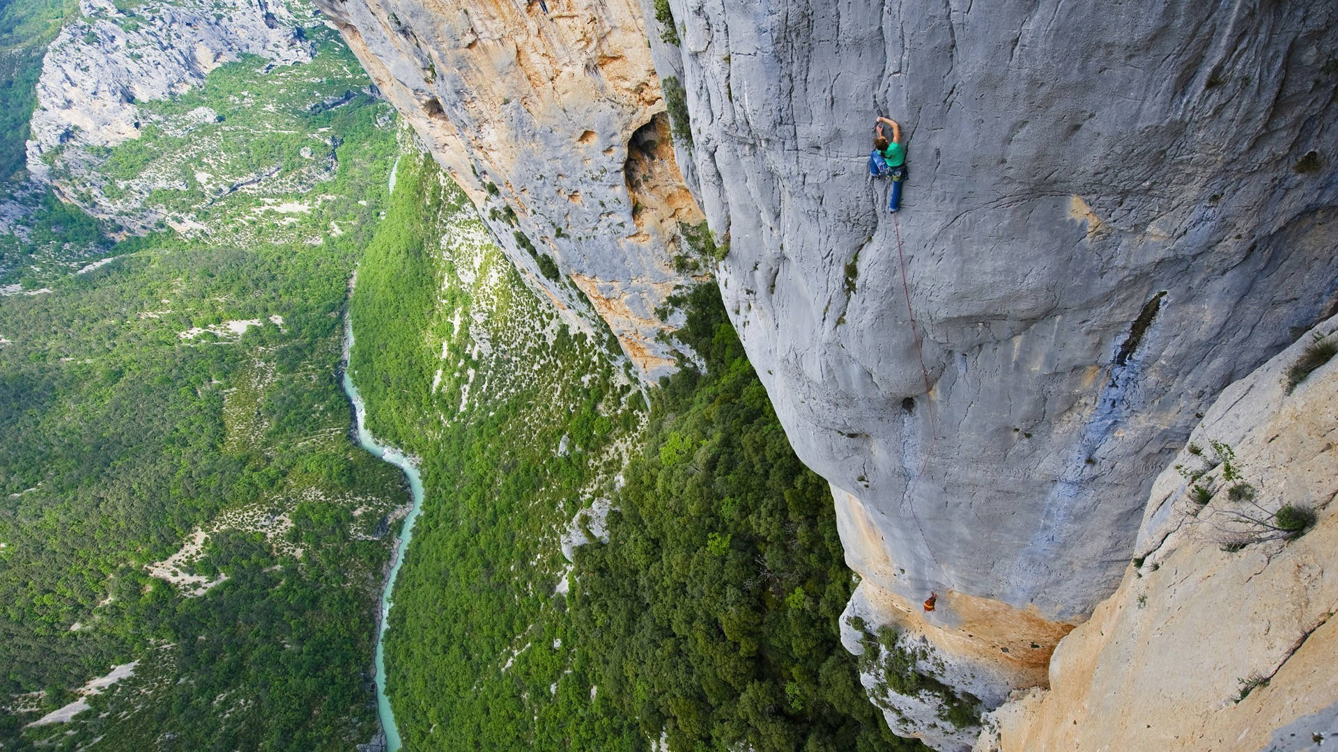 Climbing 桌布 壁紙 — 一名攀岩者攀爬陡峭的懸崖面，下方是蜿蜒穿過翠綠峽谷的河流。