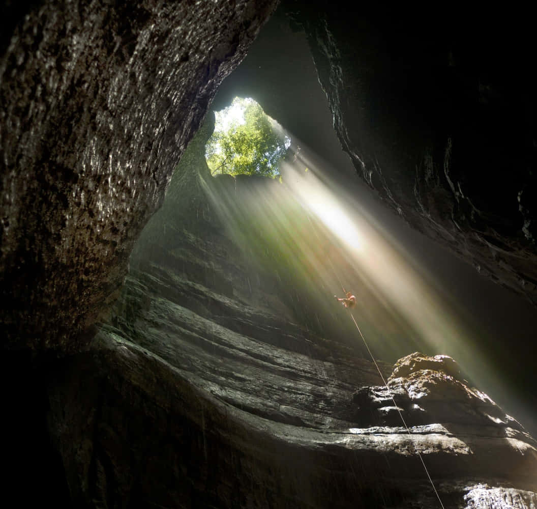 Cave 桌布 壁紙 — 一名攀岩者沿繩索下降至黑暗洞穴，上方狹窄開口處有陽光射入。