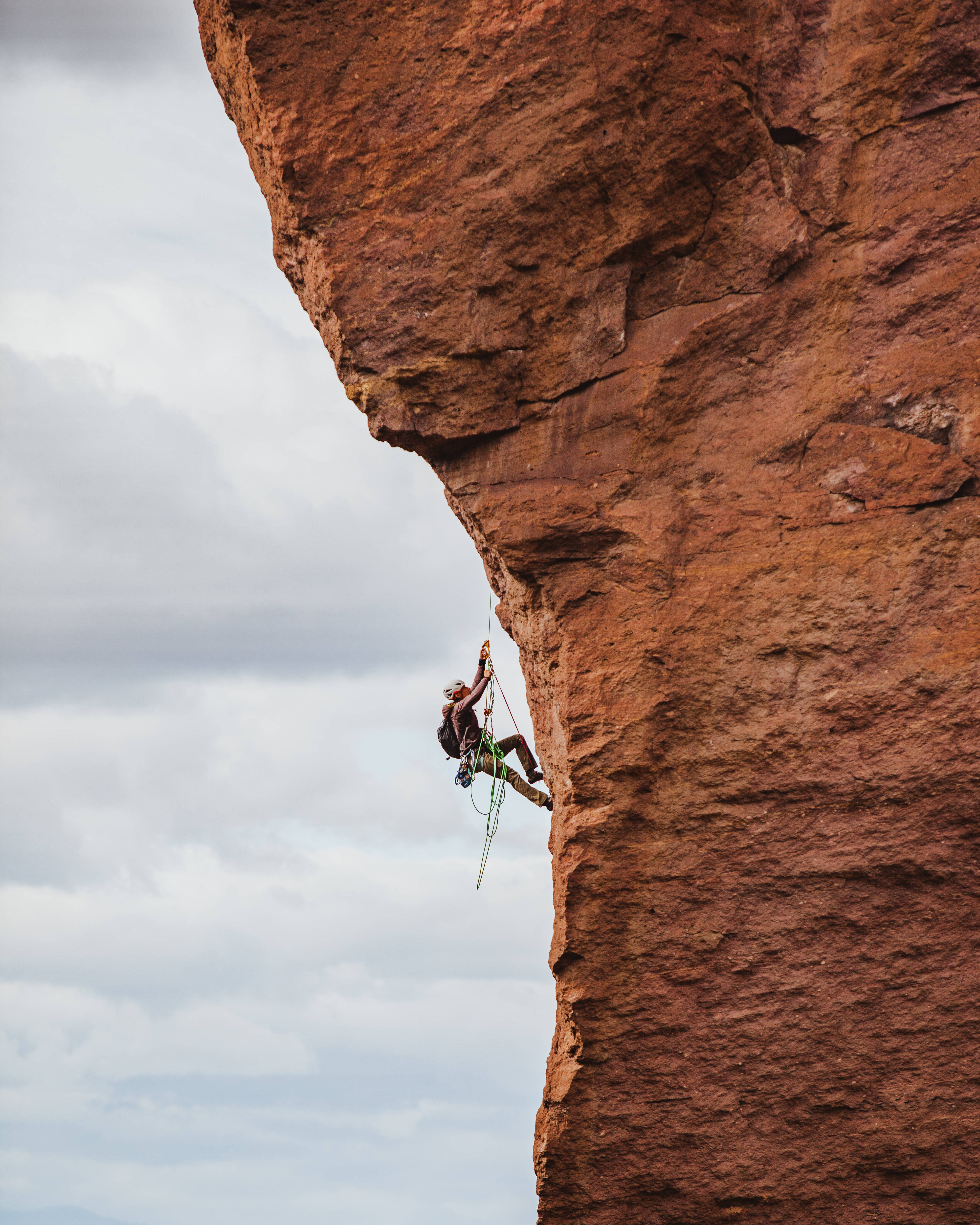 Climbing 桌布 壁紙 — 一名攀岩者用繩索和裝備攀爬陡峭的紅色岩壁，背景是多雲的天空。