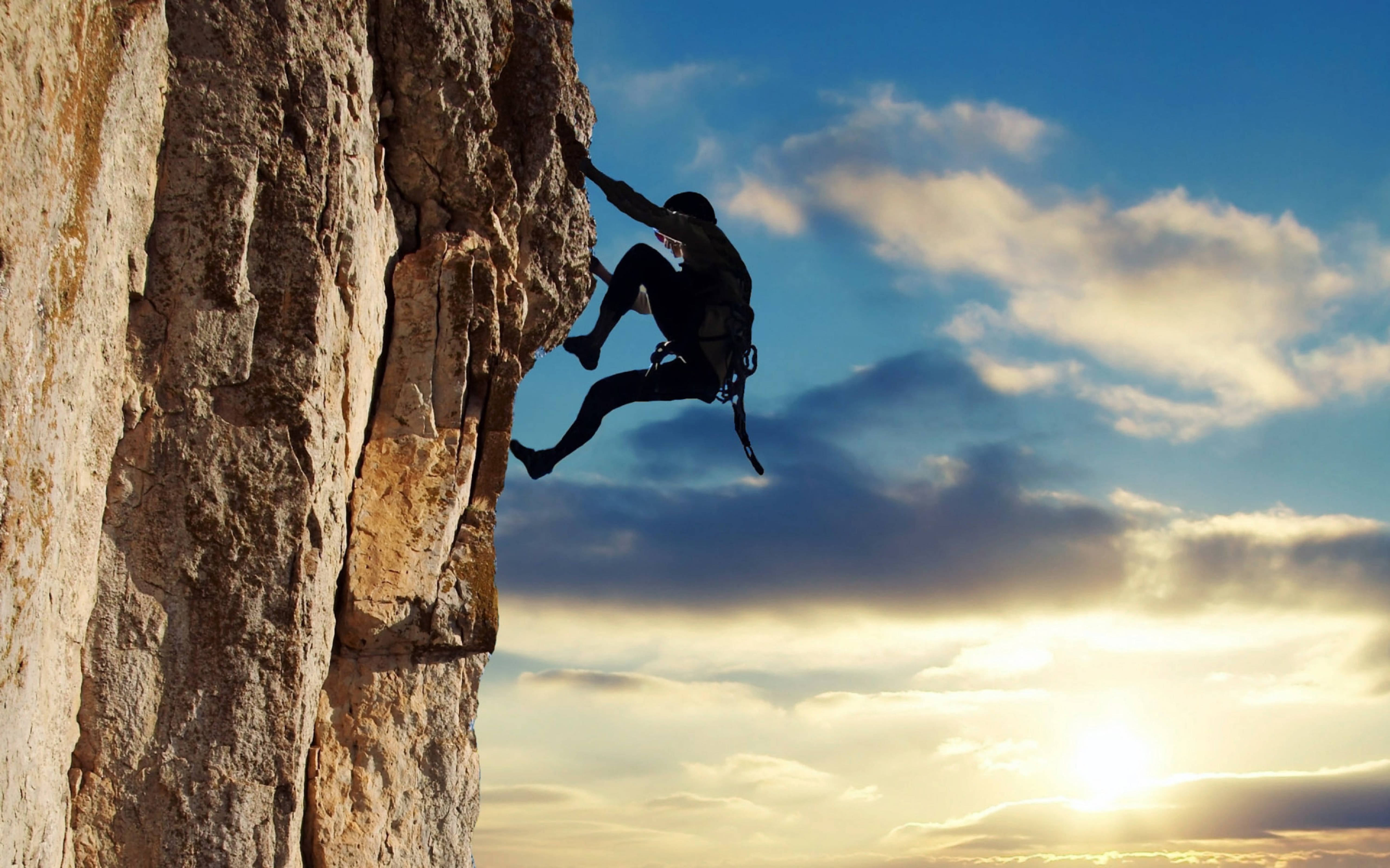 Extreme 桌布 壁紙 — 一名攀岩者的剪影在金色雲彩的戲劇性日落天空下攀登崎嶇的懸崖面。