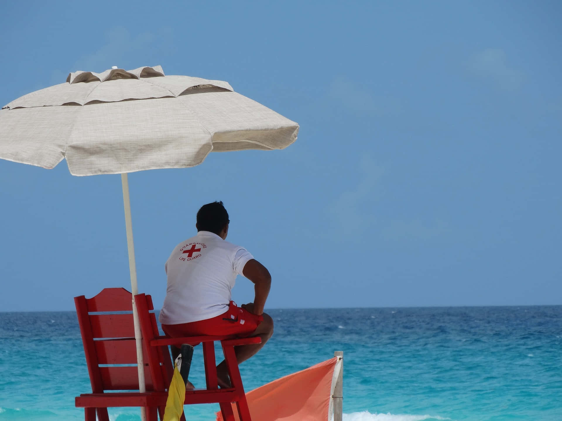 Cancun 桌布 壁紙 — 一名救生員坐在紅色椅子上，在米色遮陽傘下，眺望著清澈藍天下碧綠色的海水。