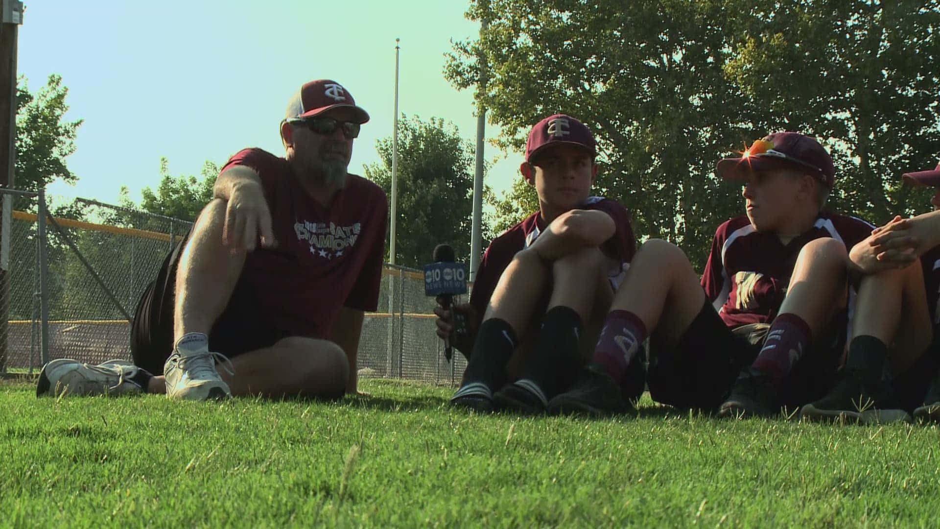Little League Baseball 桌布 壁紙 — 一名教練和穿著棗紅色球衣的年輕棒球運動員坐在陽光普照的草地上，其中一人手持麥克風。