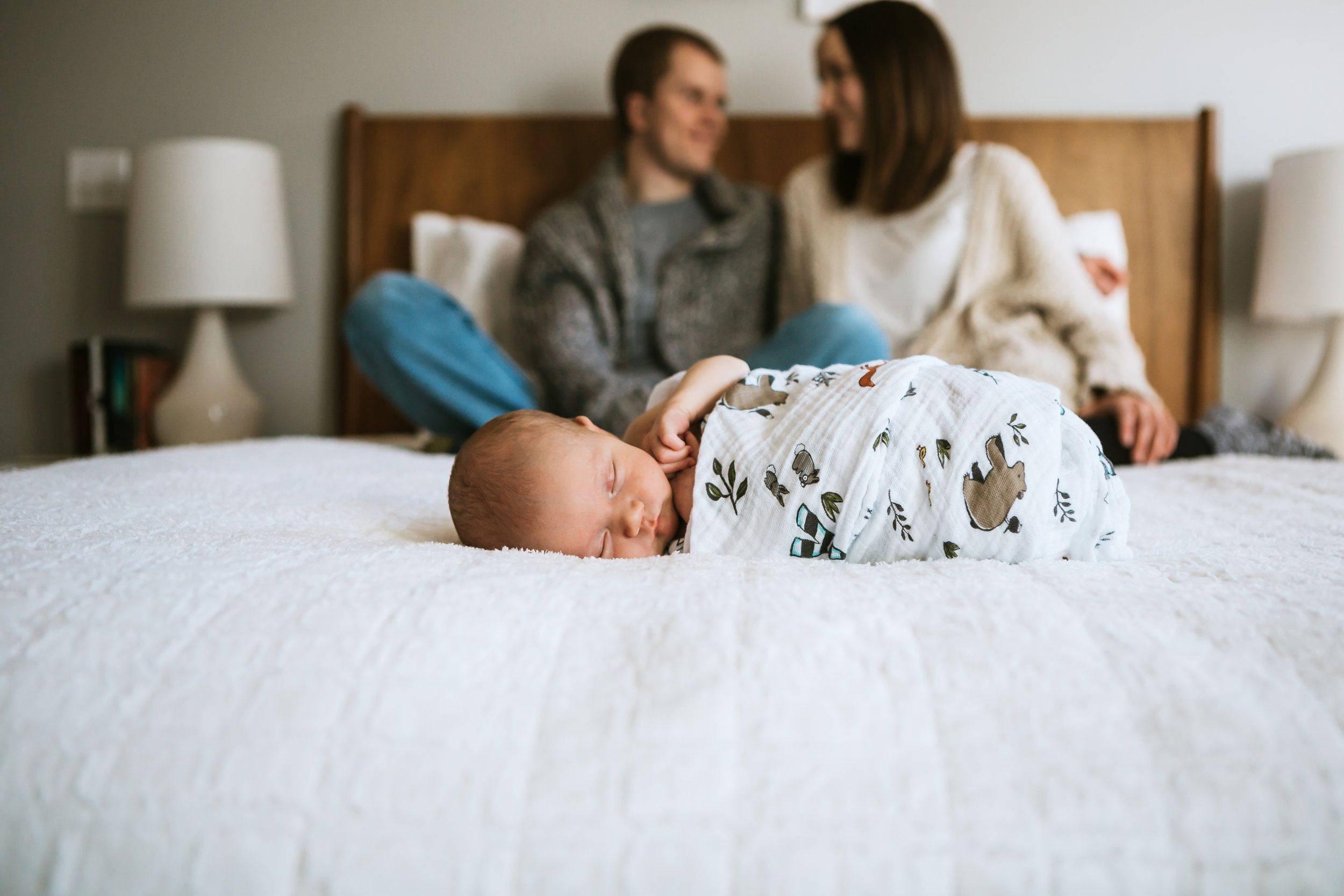 Mom 桌布 壁紙 — 一名新生嬰兒在白色紋理床上安睡，父母在背景中溫柔微笑。