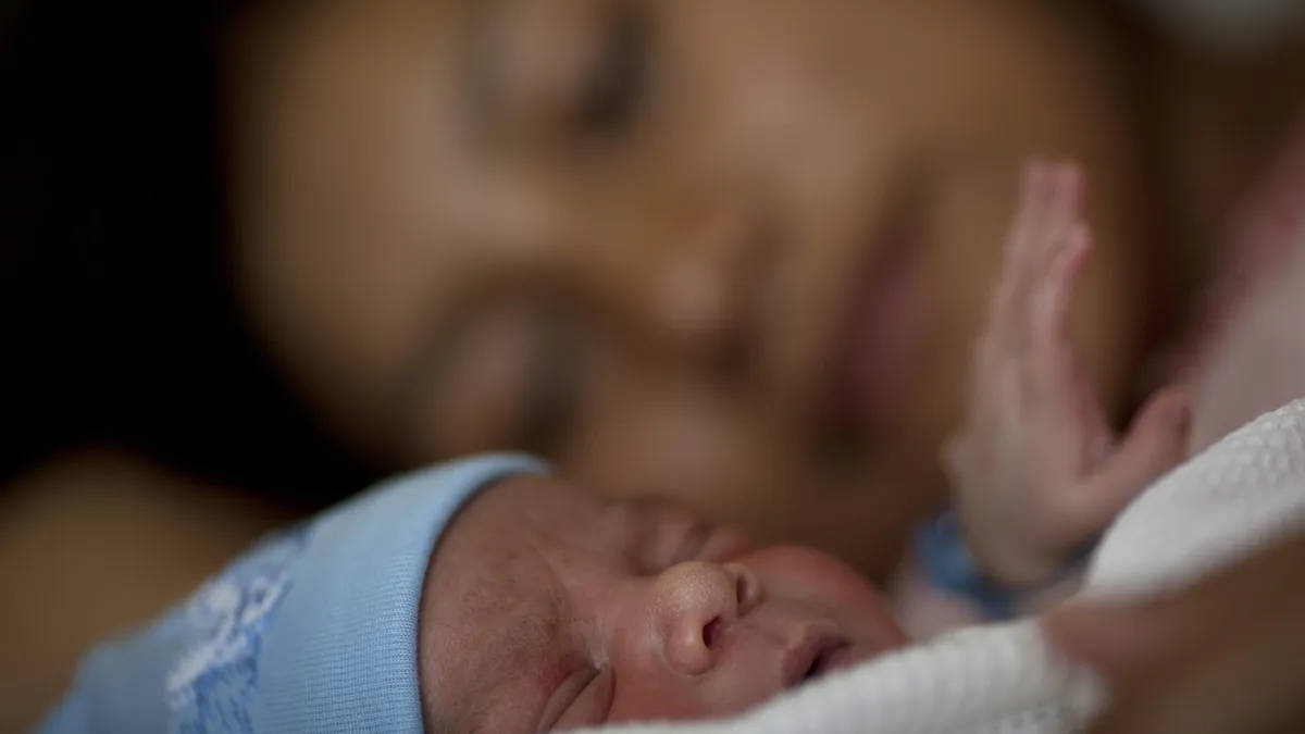 Newborn 桌布 壁紙 — 一名新生嬰兒戴著藍色帽子安詳入睡，背景中模糊的母親深情地凝視著。
