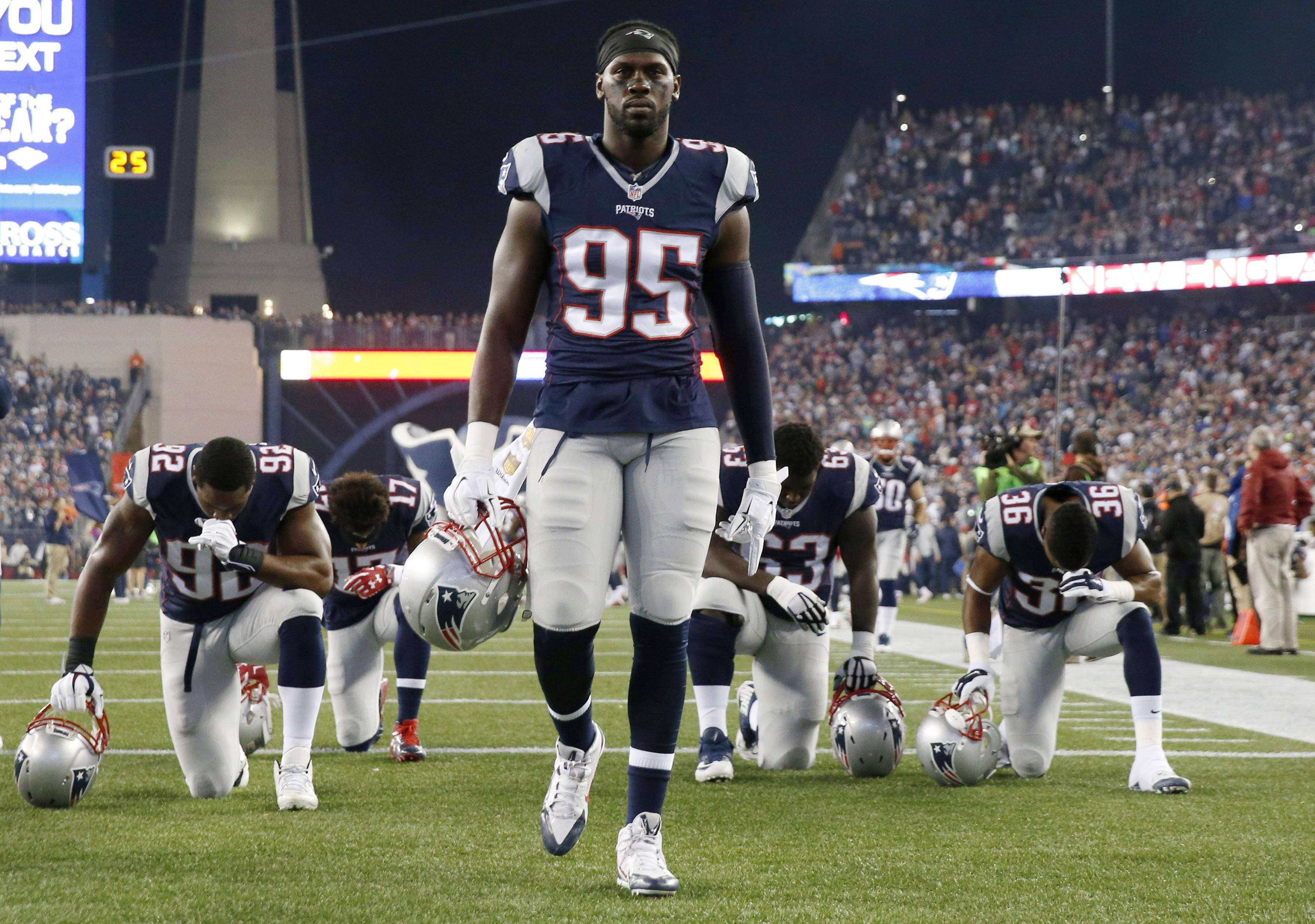 New England Patriots 桌布 壁紙 — 一名新英格蘭愛國者隊的美式足球員在球場燈光下莊嚴地走過球場，而隊友則跪地抗議。