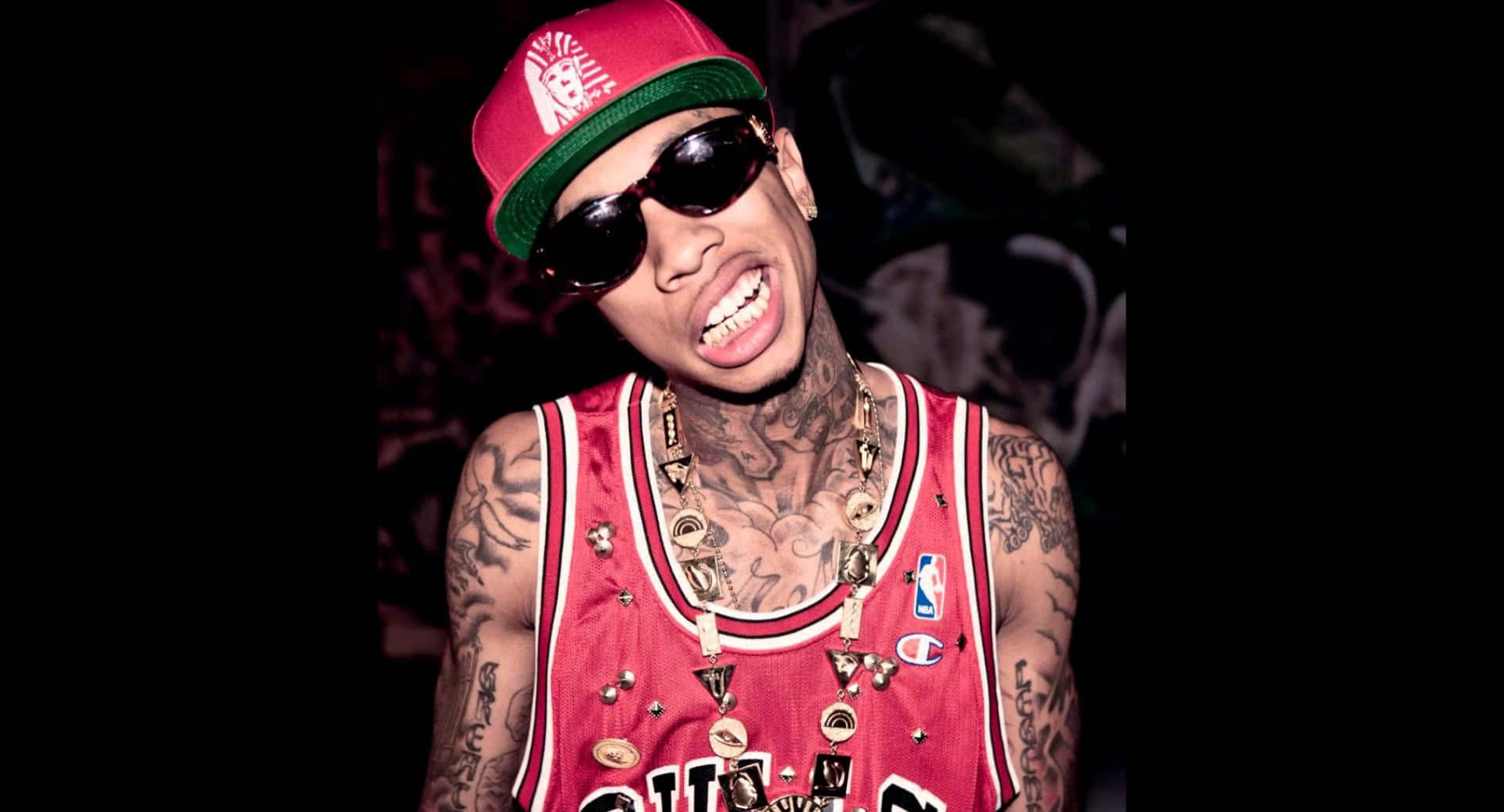 Tyga 桌布 壁紙 — 一名有刺青和戴太陽眼鏡的男子，穿著紅色籃球球衣和帽子，在深色背景下咧嘴大笑。