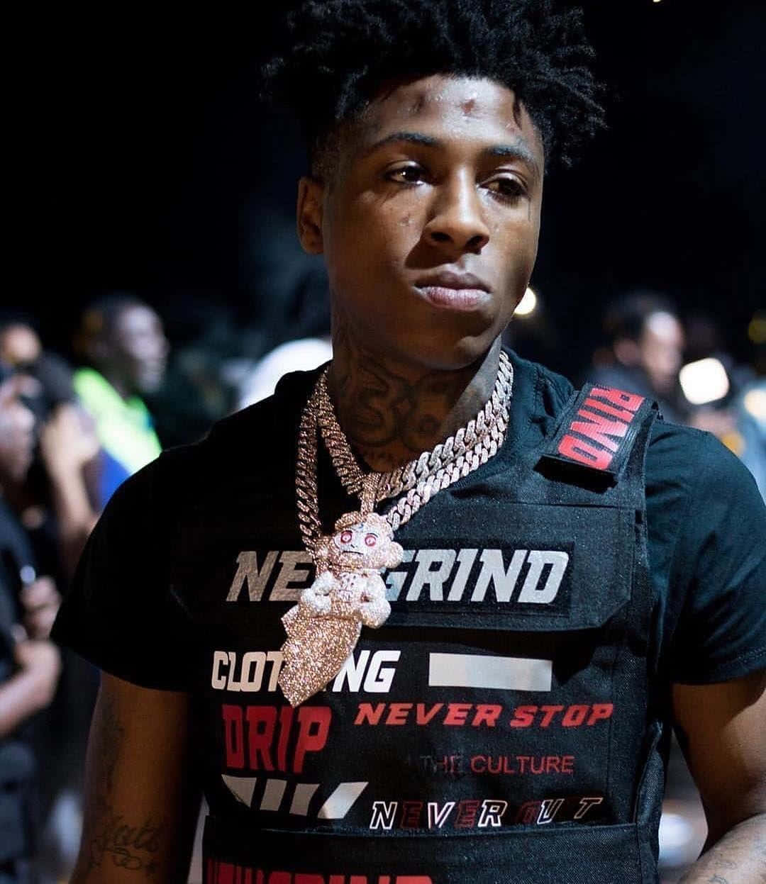Youngboy 桌布 壁紙 — 一名有刺青和戴金鍊的年輕男子，在舞台燈光下穿著黑色圖案背心，背後是模糊的人群。