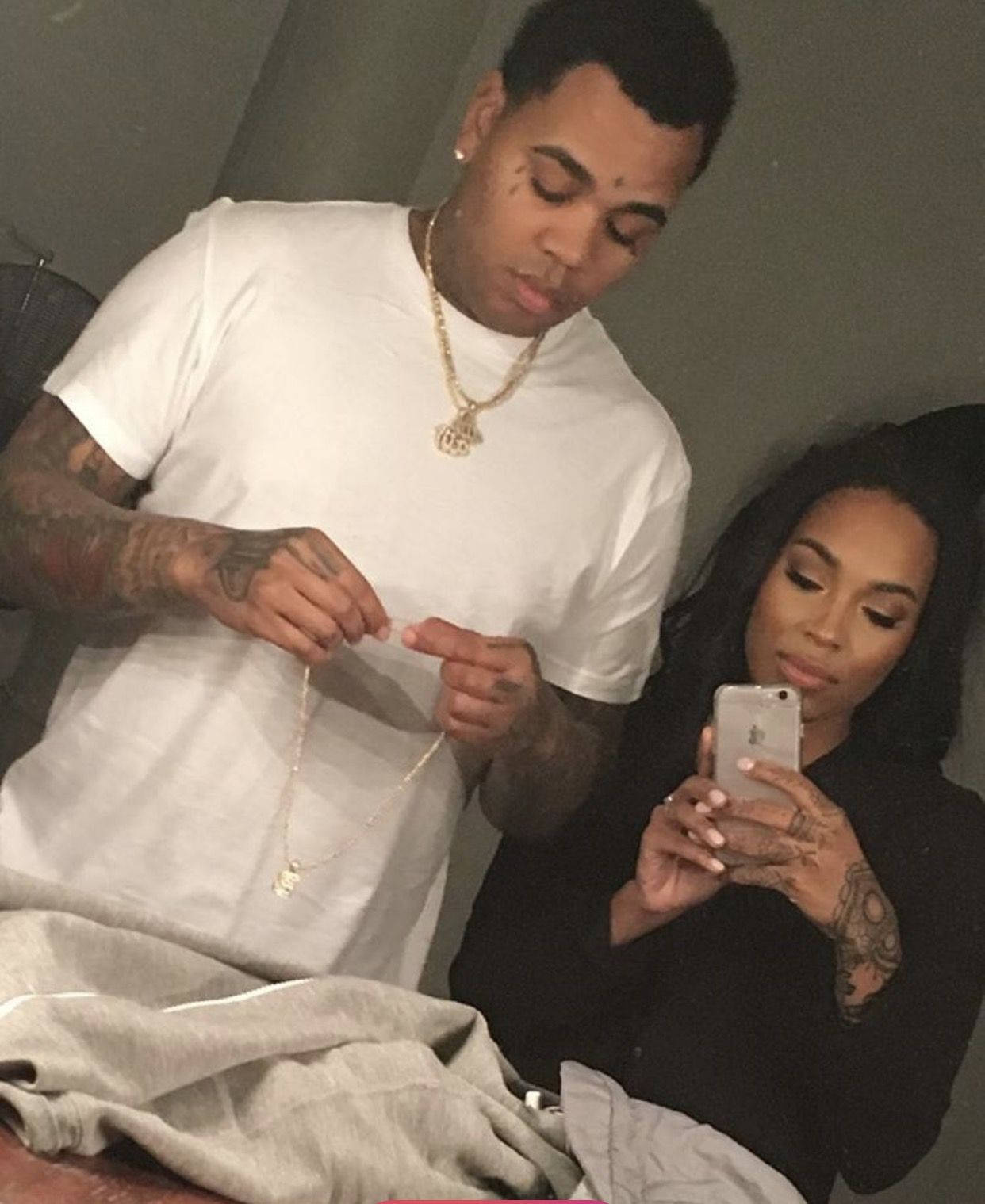 Kevin Gates 桌布 壁紙 — 一名有刺青和戴金鍊的男人拿著一條項鍊，而一名穿黑衣的女人在昏暗的房間裡用手機自拍。