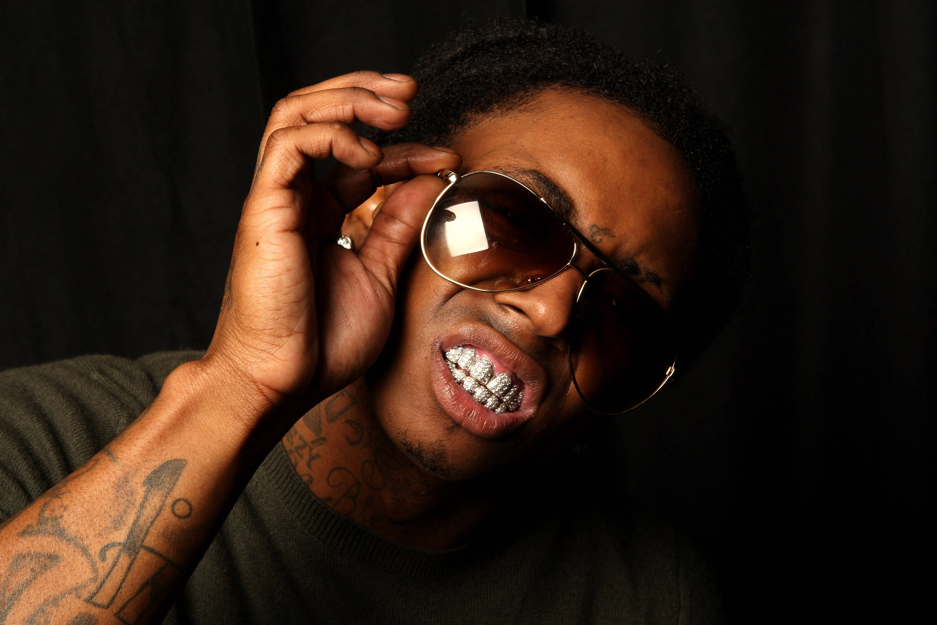 Lil Wayne 桌布 壁紙 — 一名有刺青和金色牙套的男子在黑色背景下調整太陽眼鏡。