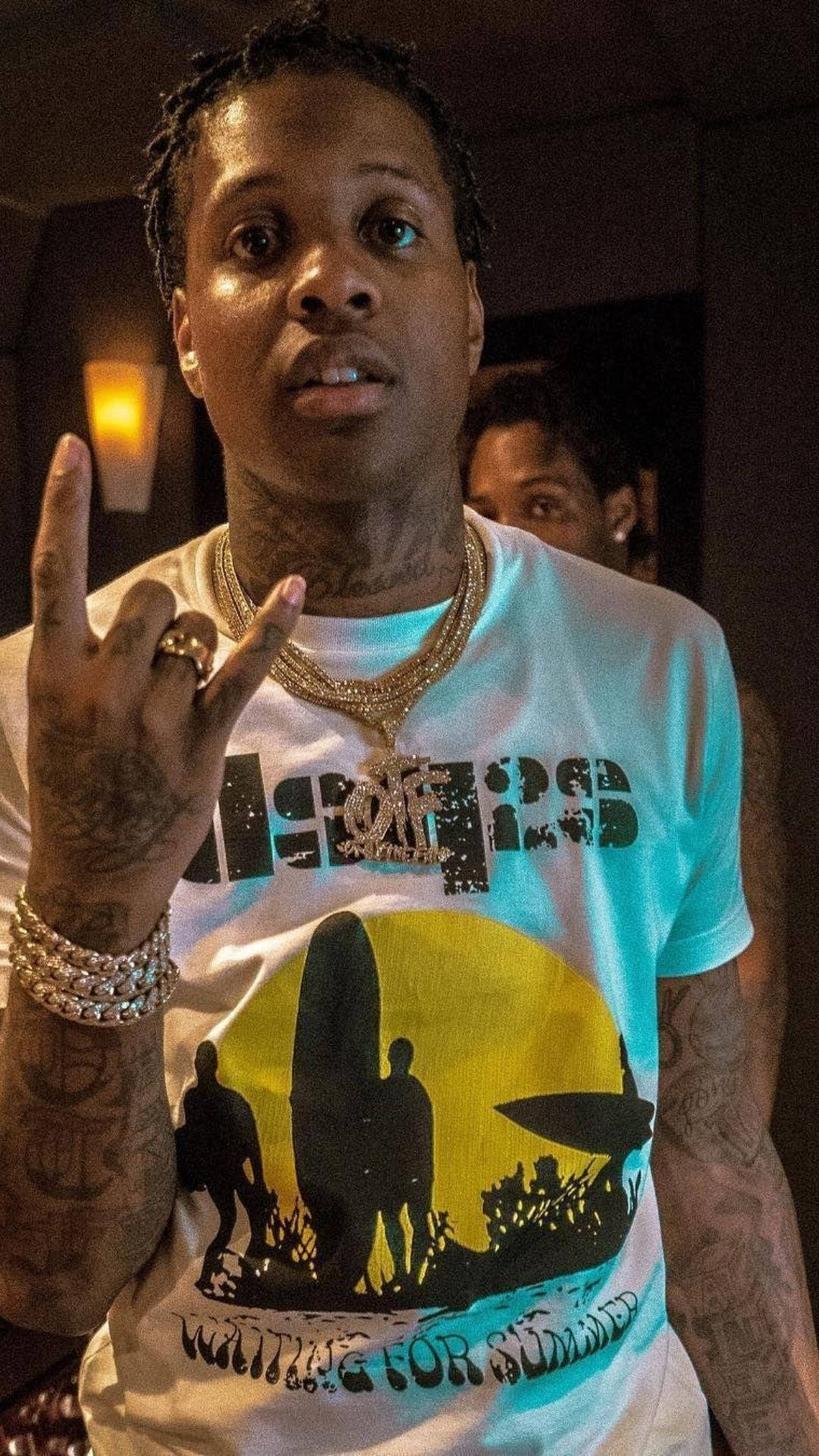 Lil Durk 桌布 壁紙 — 一名有刺青和黃金首飾的男子在昏暗的房間裡穿著圖案T恤做手勢。