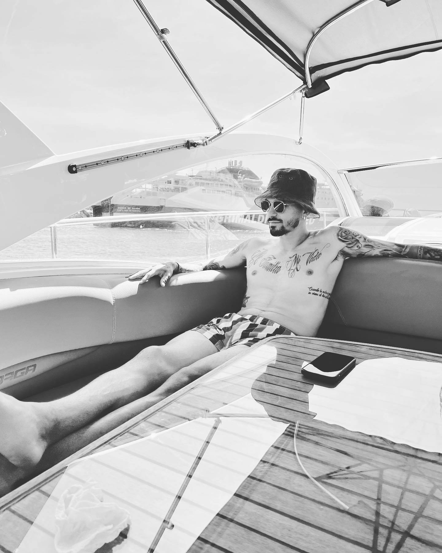 Yacht 桌布 壁紙 — 一名有刺青的男士在遊艇甲板上，在遮陽篷下赤裸上身休息，背景可見一艘郵輪。