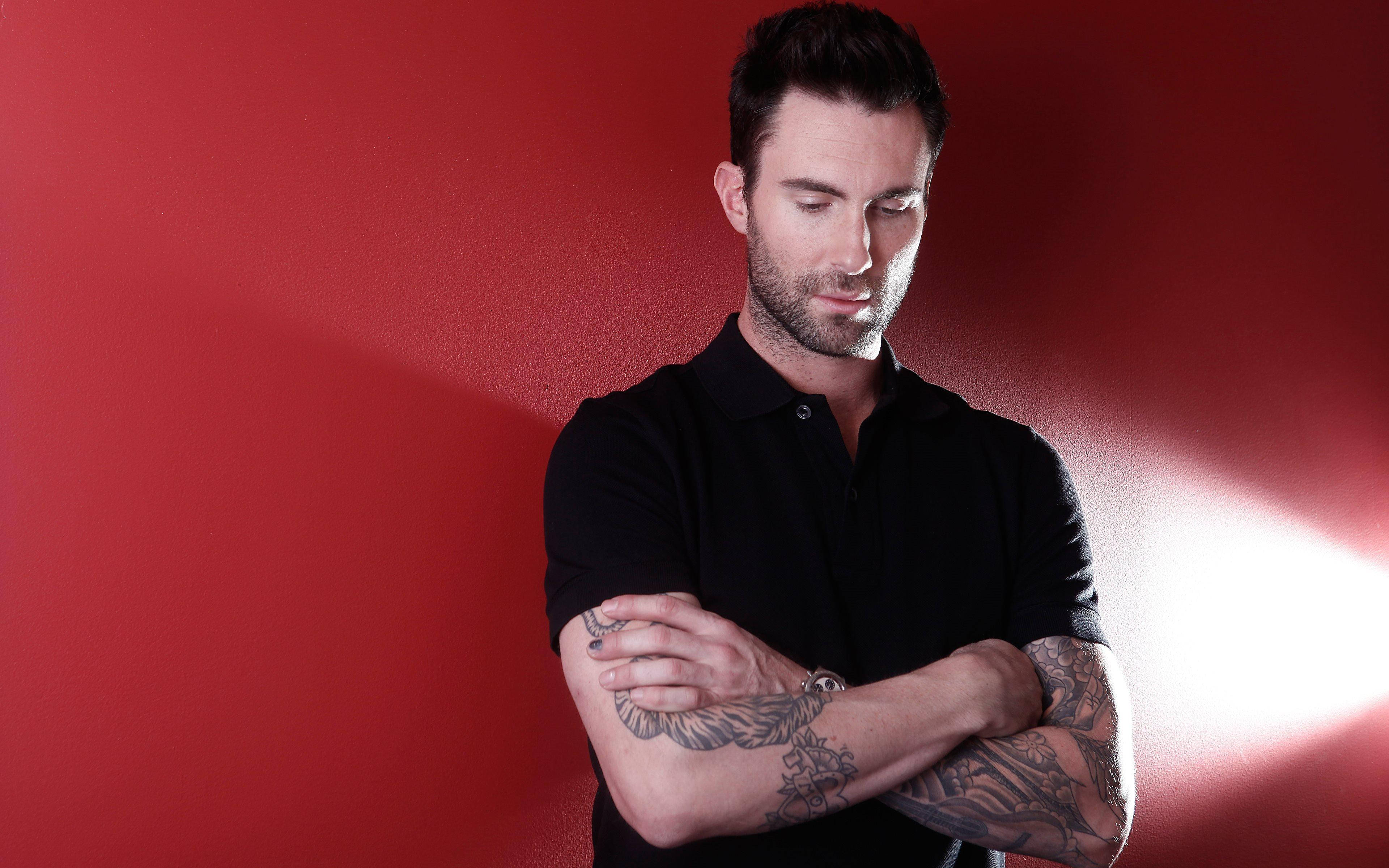 Adam Levine 桌布 壁紙 — 一名有刺青的男子靠著鮮紅色的牆壁，雙臂交叉，被聚光燈照亮。