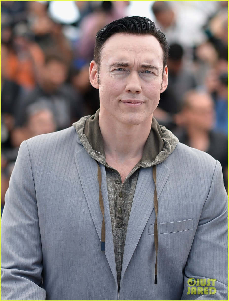 Kevin Durand 桌布 壁紙 — 一名梳著向後梳的頭髮的男子，在模糊的人群前穿著灰色西裝外套在連帽衫外面。