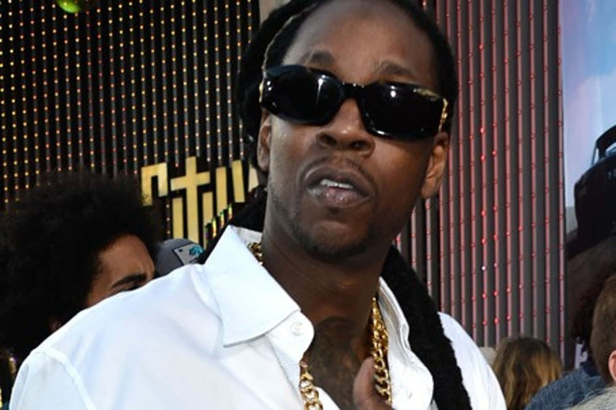 2 Chainz 桌布 壁紙 — 一名梳辮子、戴太陽眼鏡的男子，身穿白襯衫和金色項鍊，背景閃爍。