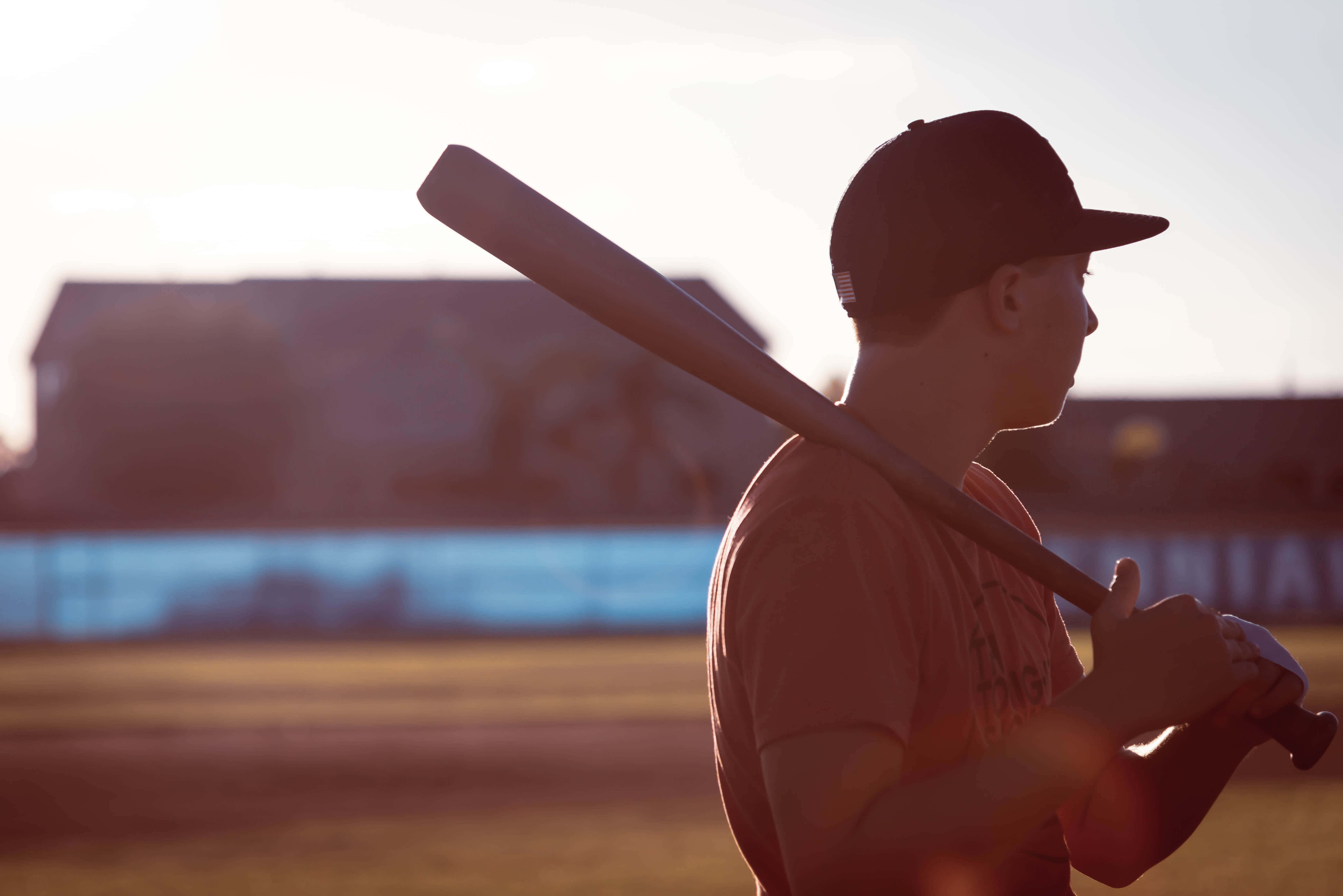 baseball bat 桌布 壁紙 — 一名棒球運動員的剪影在日落時分的球場上，手持球棒，沐浴在溫暖的金色光芒中。