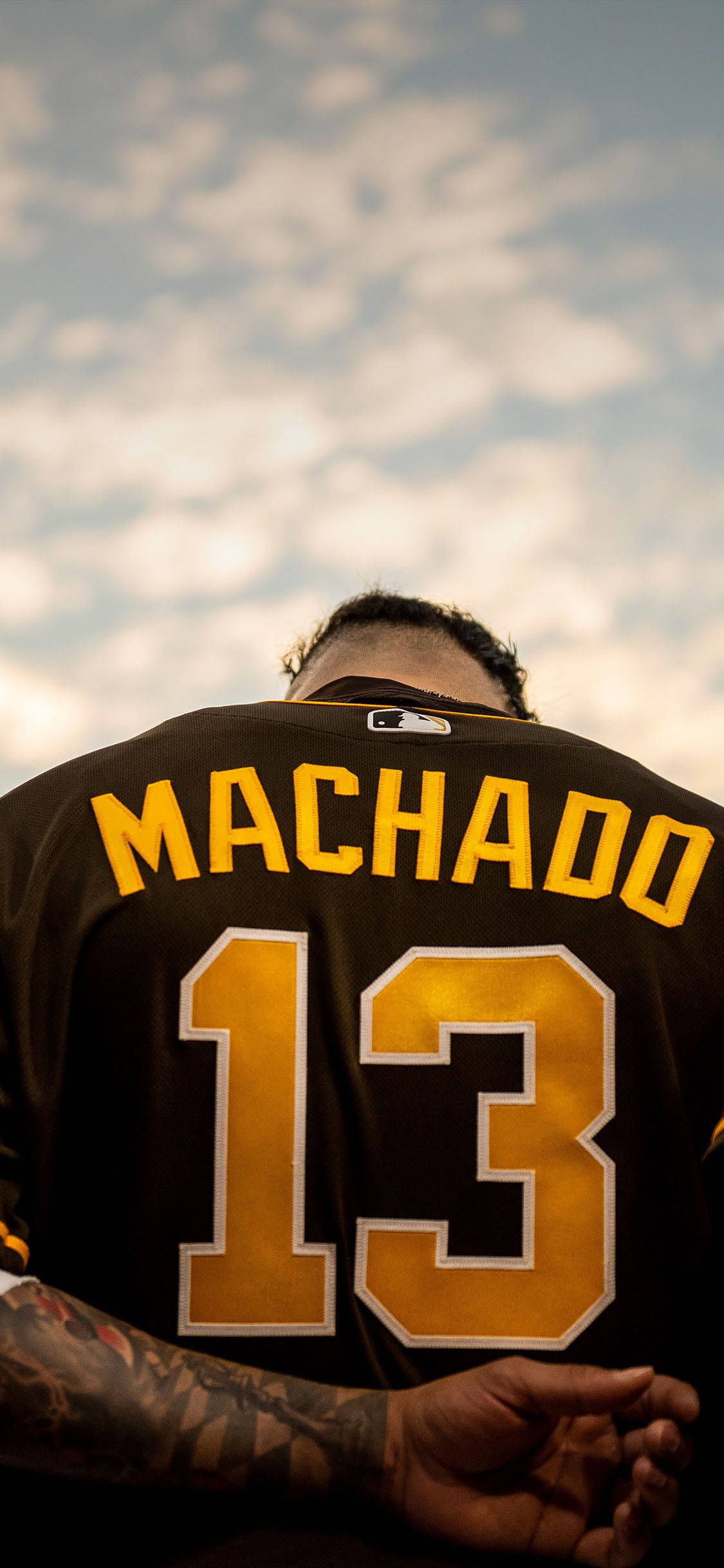 Machado 桌布 壁紙 — 一名棒球運動員身穿黑金色 Machado 13 球衣，背對多雲的天空。