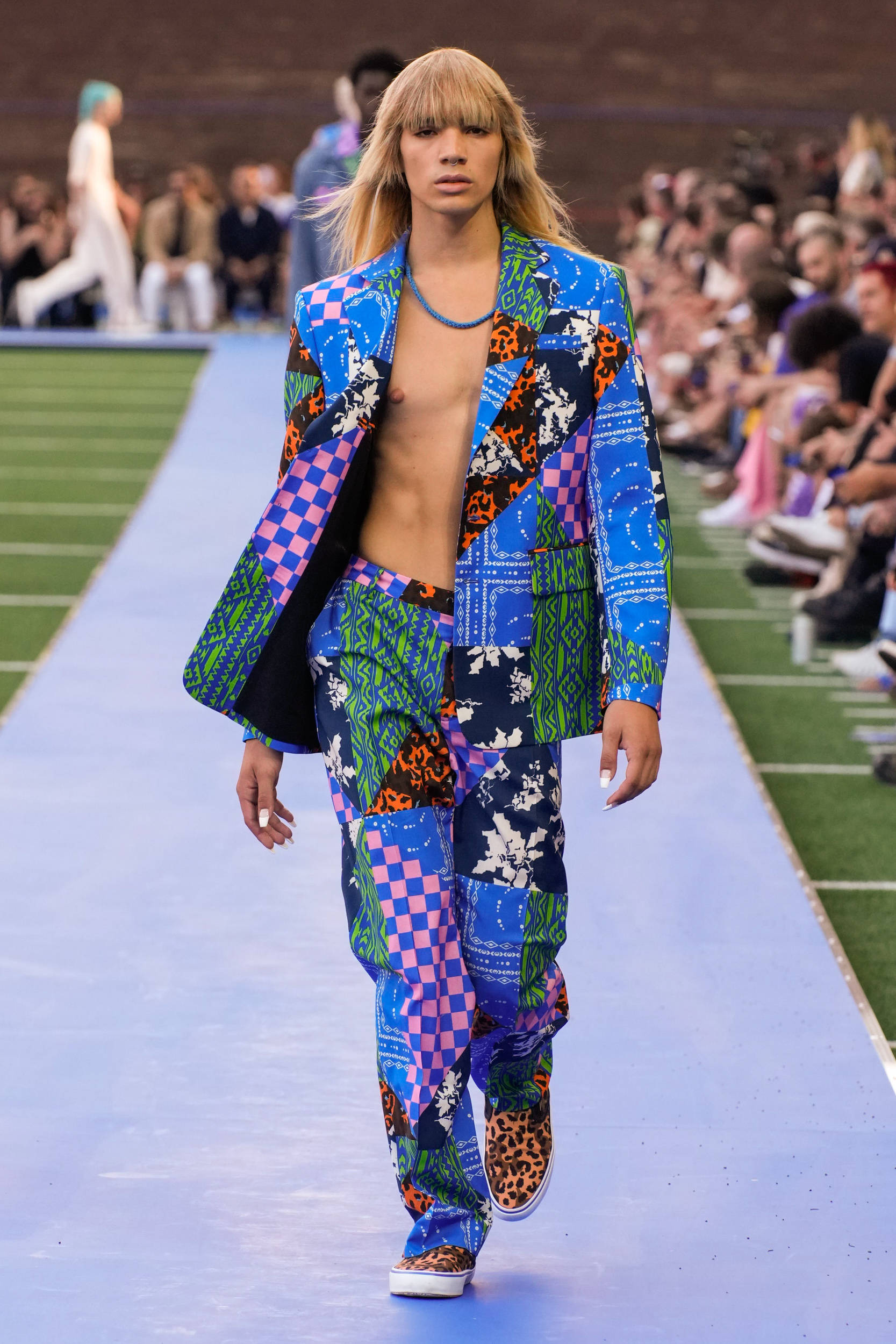 Fashion Model 桌布 壁紙 — 一名模特兒在藍色伸展台上走秀，身穿鮮豔的拼布套裝，帶有豹紋和幾何圖案。