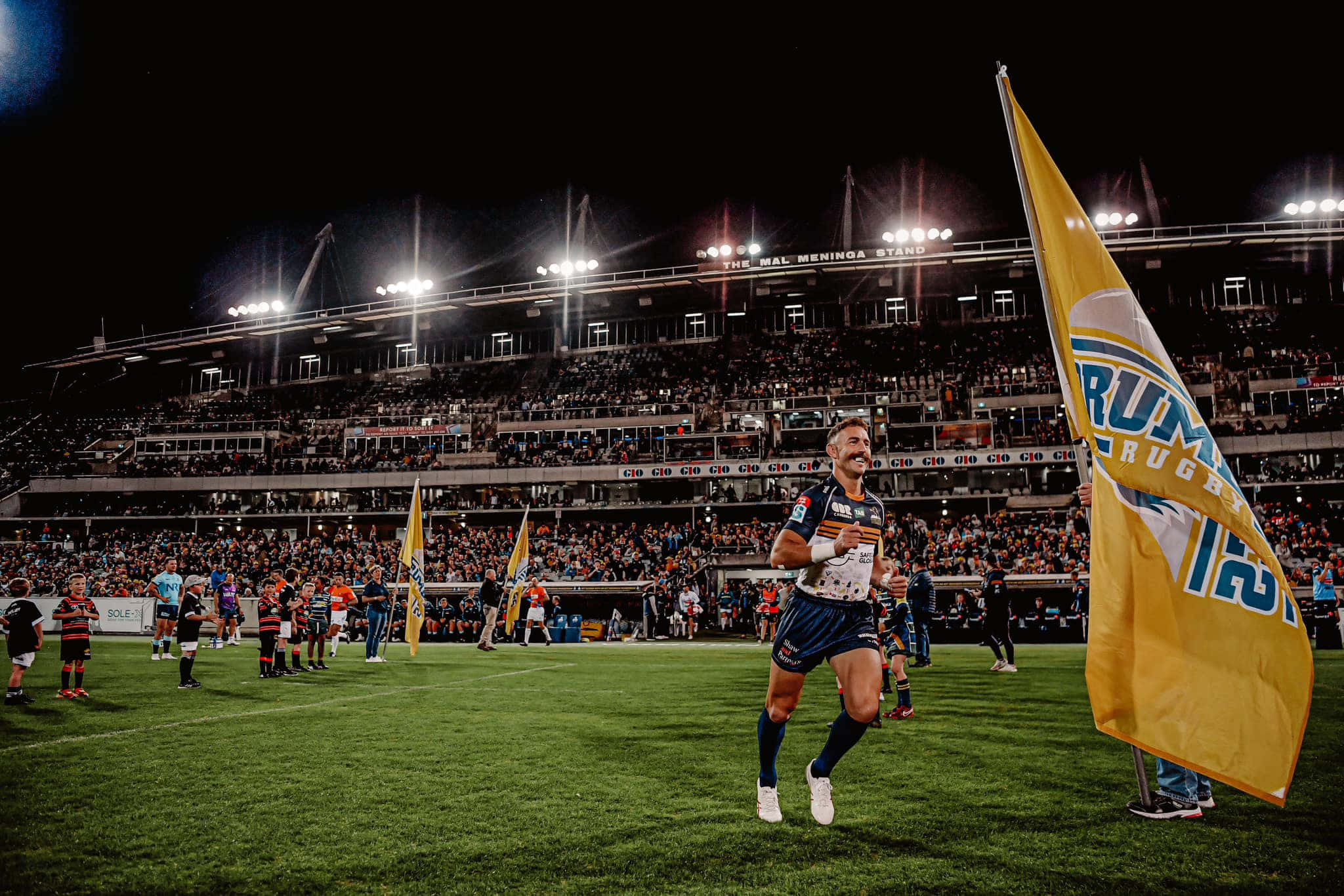 Brumbies 桌布 壁紙 — 一名橄欖球運動員在夜間球場燈光下慶祝勝利，看台上球迷揮舞旗幟。