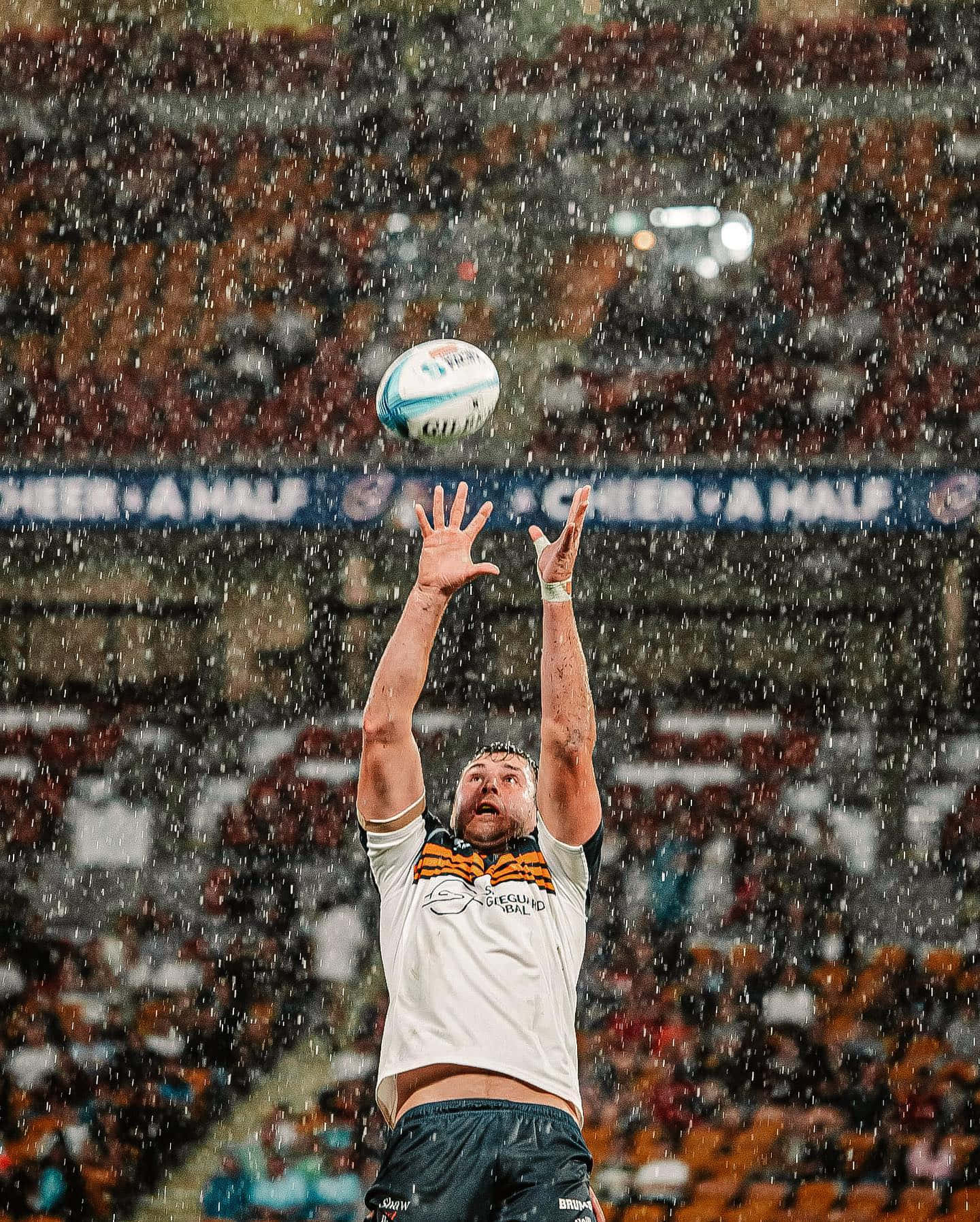 Brumbies 桌布 壁紙 — 一名橄欖球運動員在雨中跳起，在球場燈光下伸手接球，身後是擠滿的人群。