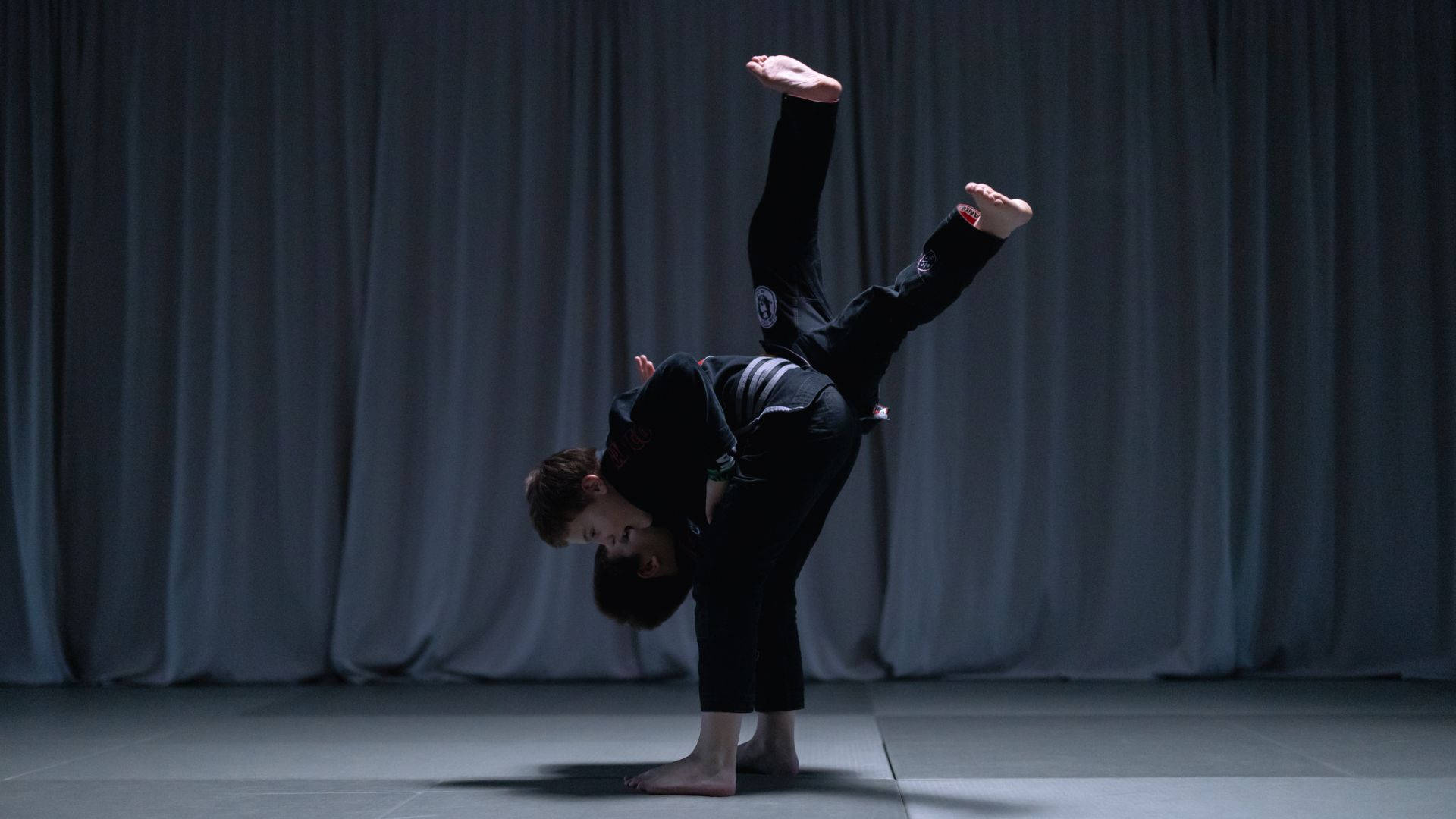 martial arts 桌布 壁紙 — 一名武術家在黑暗帷幕前，於戲劇性聚光燈下在墊子上做出動態的倒立劈腿動作。