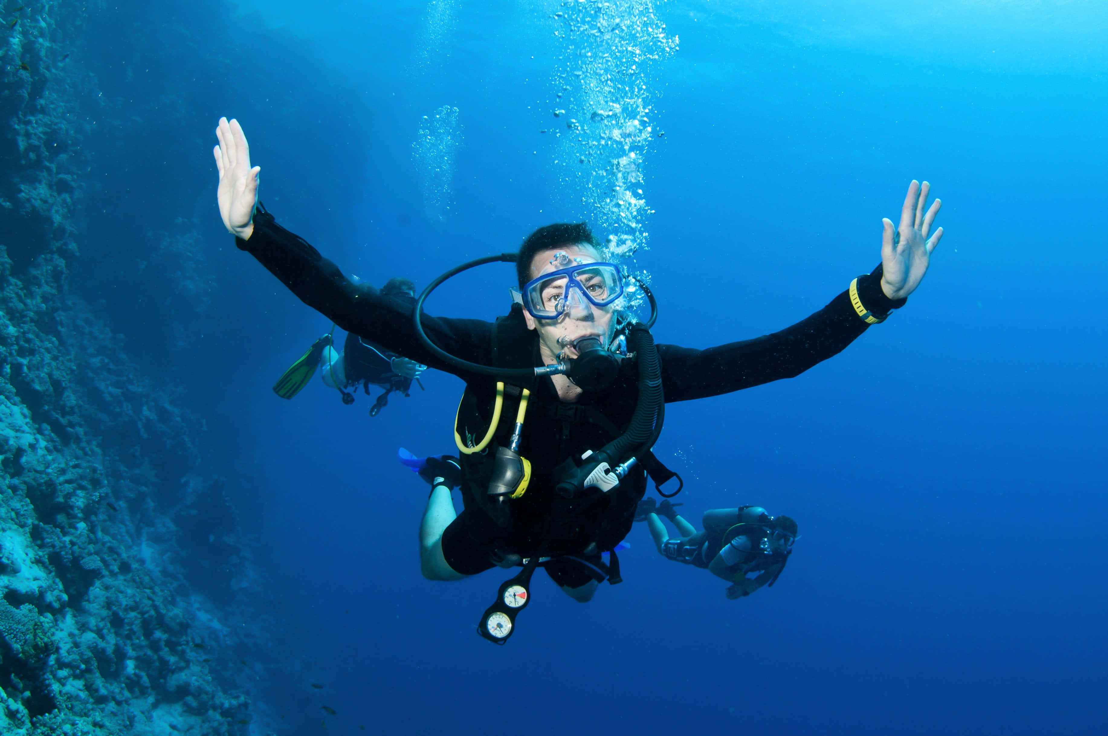 diving 桌布 壁紙 — 一名水肺潛水員張開雙臂，在珊瑚礁附近的水中快樂地微笑，氣泡在他們周圍升起。