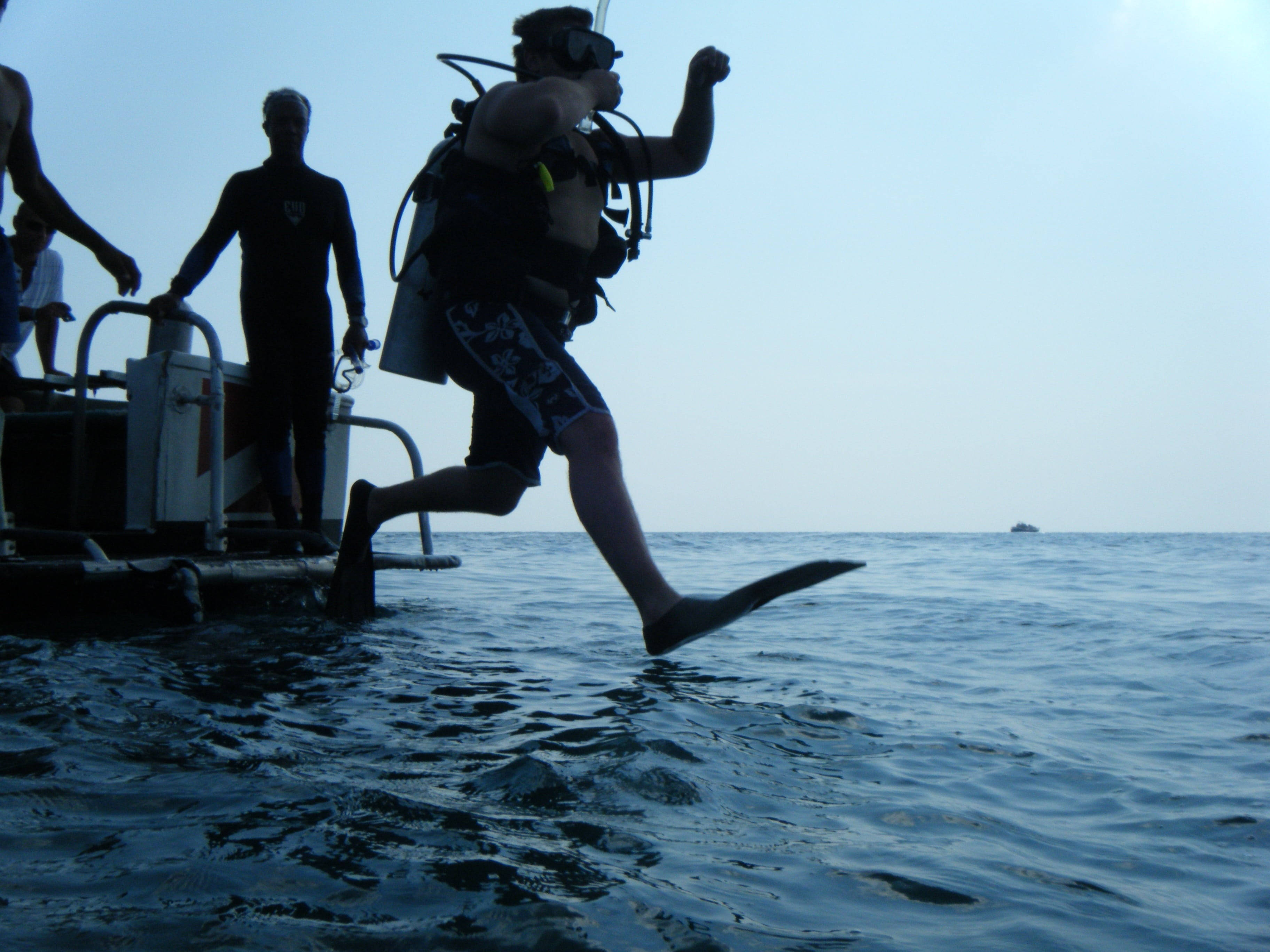 diving 桌布 壁紙 — 一名水肺潛水員從船上跳入海洋，腳蹼和裝備在明亮的天空下形成剪影。