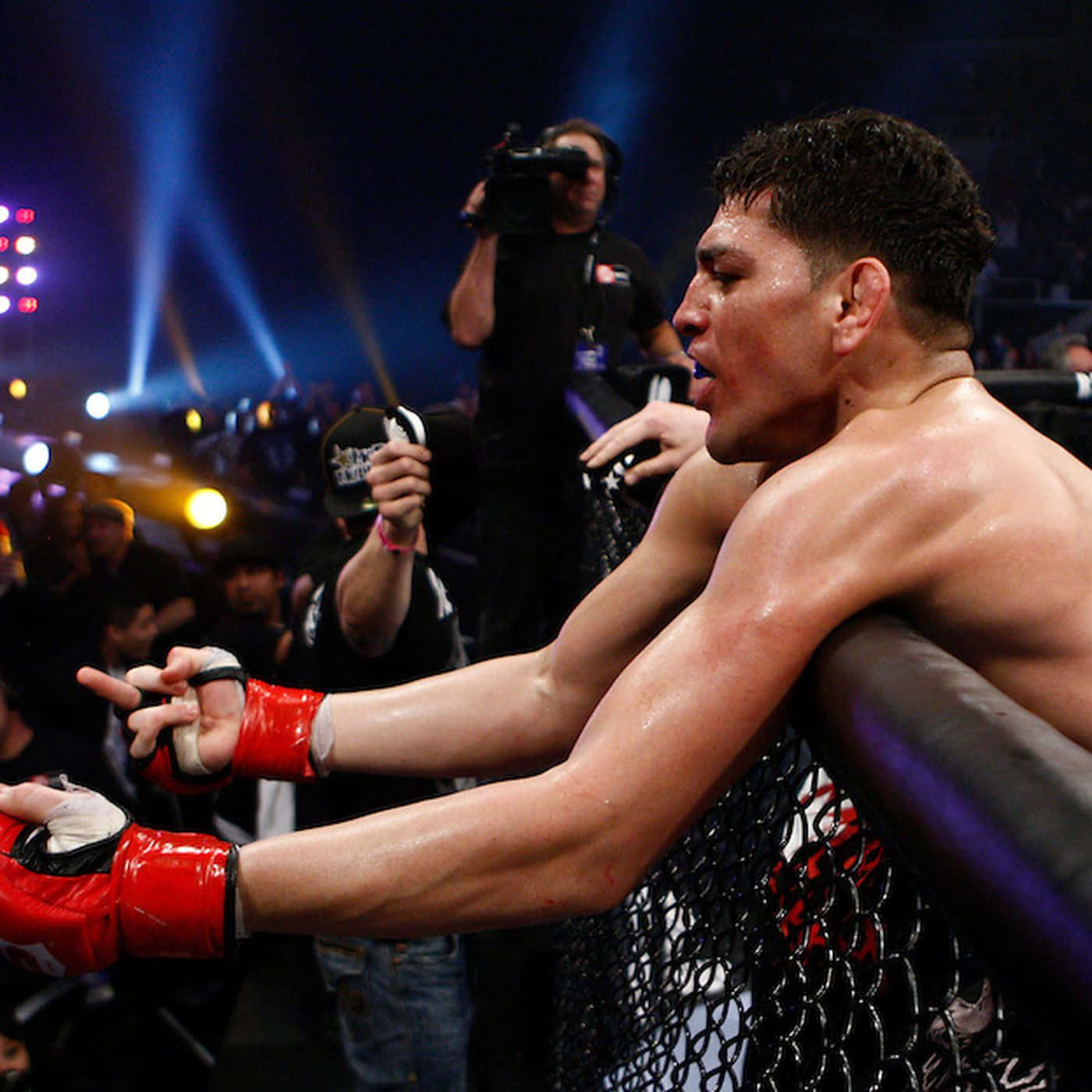 UFC 桌布 壁紙 — 一名汗流浹背、戴紅色手套的MMA拳手在昏暗的競技場中，藍色聚光燈和歡呼的人群中靠在籠邊欄杆上。