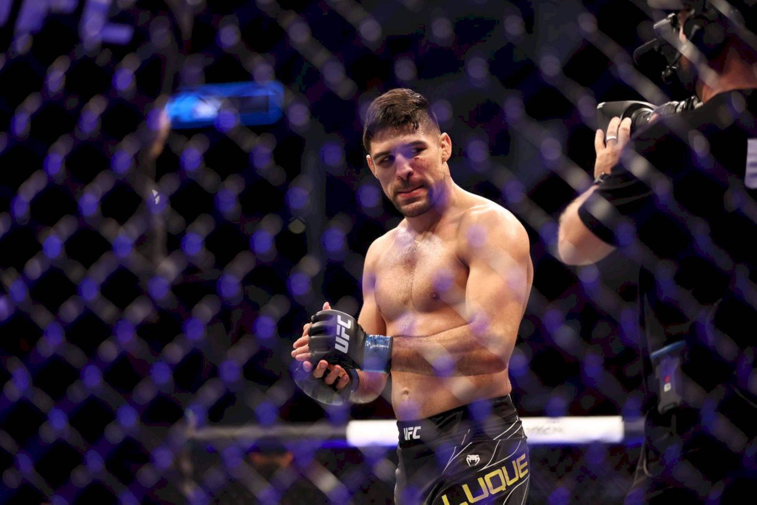 UFC 桌布 壁紙 — 一名汗流浹背的UFC拳手在明亮的場館燈光下，帶著專注的表情，手持拳套站在八角籠內。