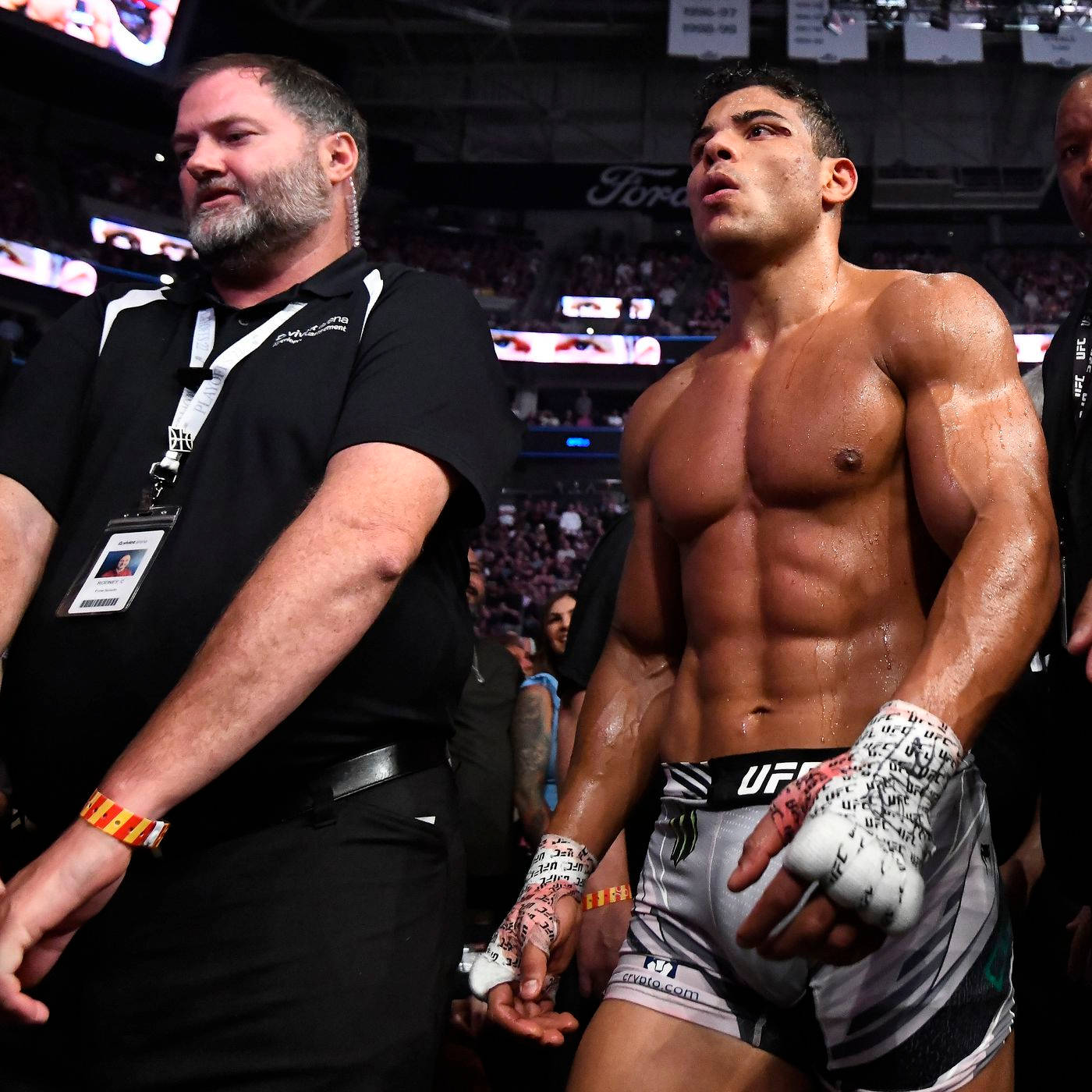 UFC 桌布 壁紙 — 一名汗流浹背的UFC拳手穿著品牌短褲和纏手帶，在明亮的燈光下與教練一起穿過擁擠的競技場走出來。