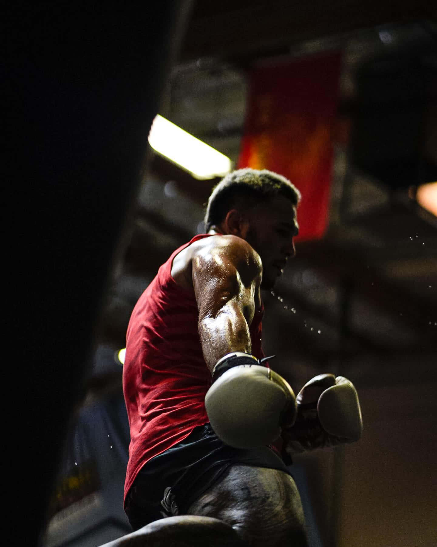 Boxing 桌布 壁紙 — 一名汗流浹背的拳擊手穿著紅色背心，在昏暗的健身房裡，在刺眼的螢光燈下打出強力一拳。