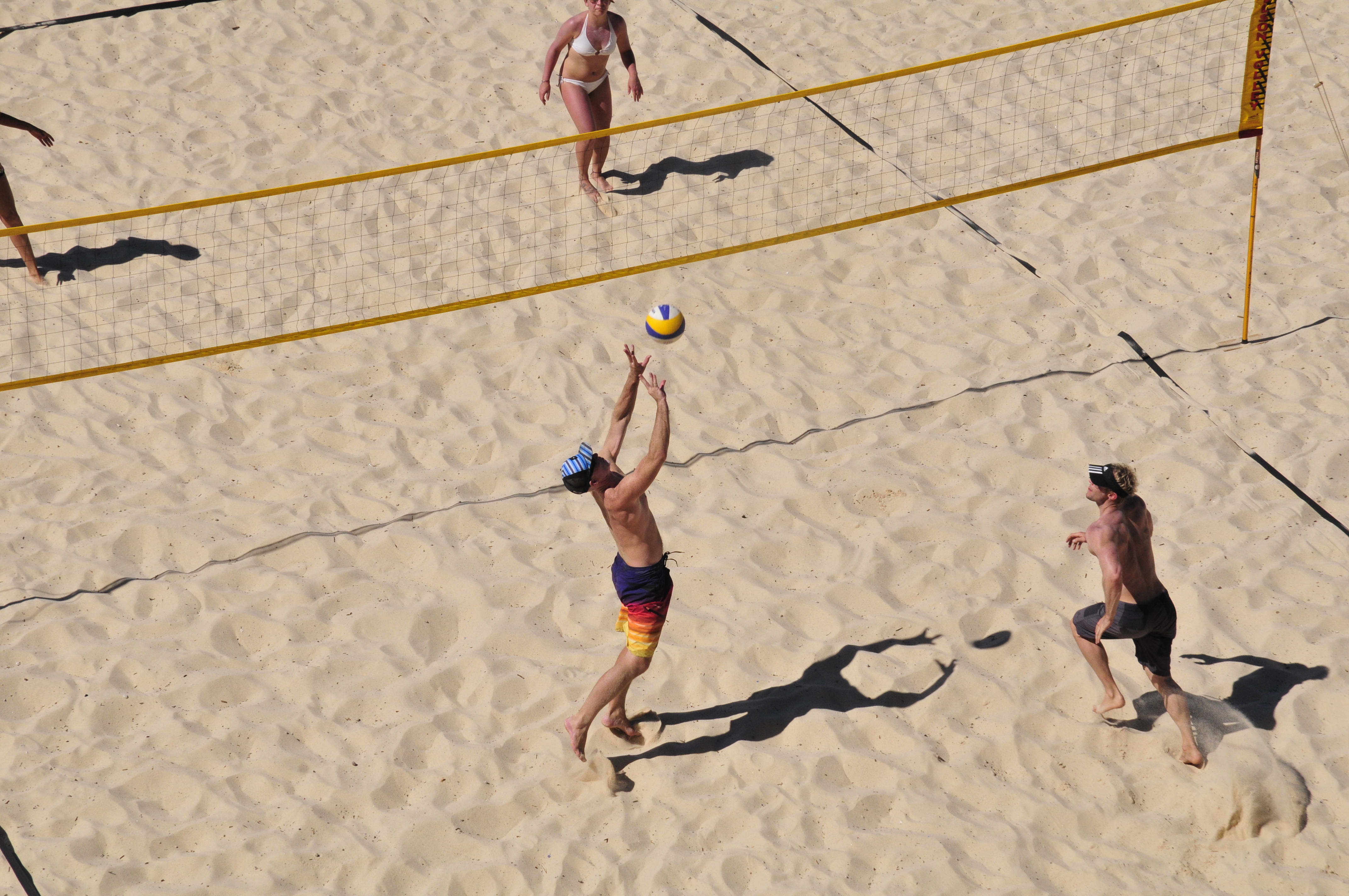 volleyball 4K 桌布 壁紙 — 一名沙灘排球運動員跳起扣球，隊友和對手在金色沙灘上、烈日下觀看。