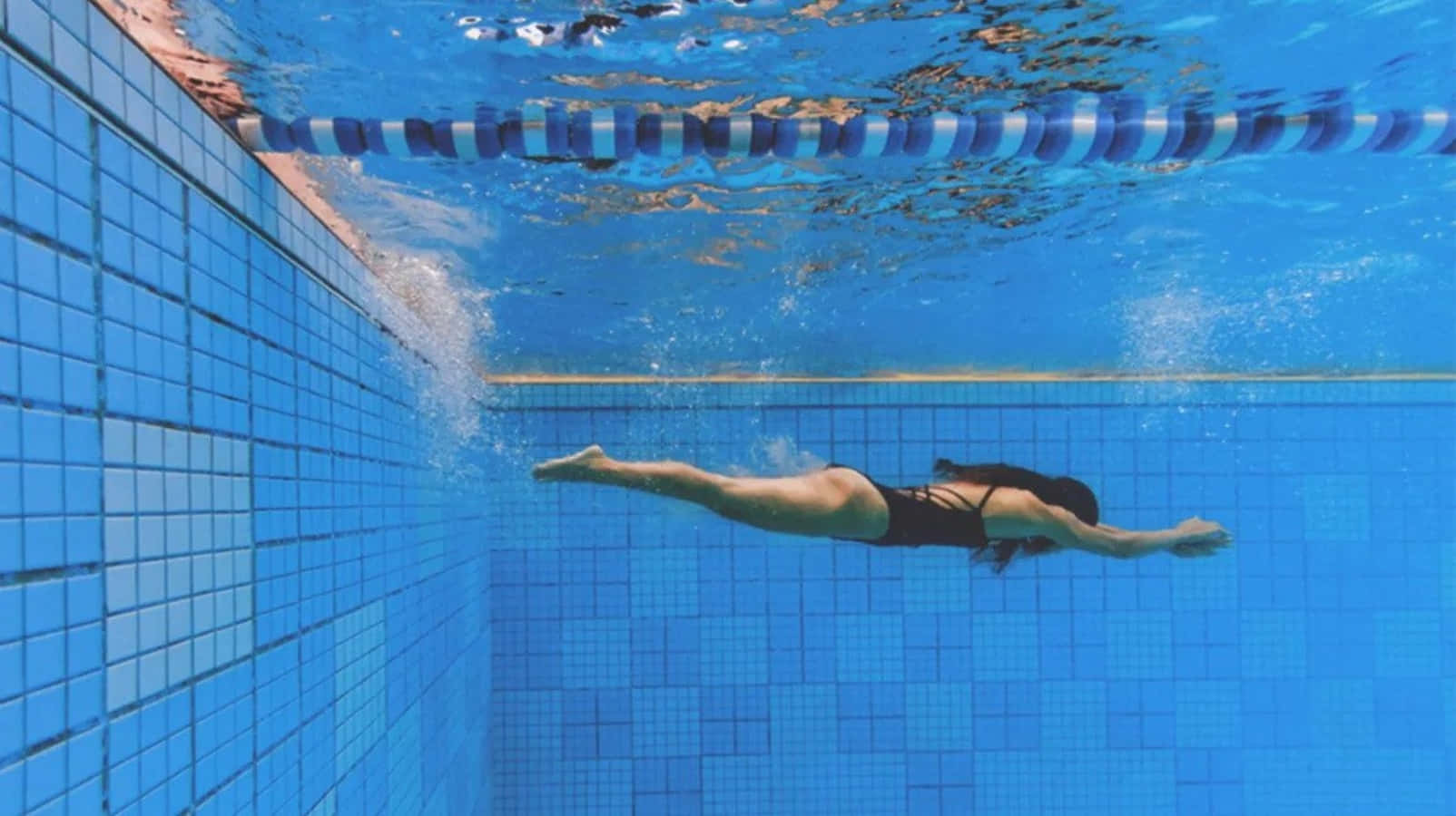 Swimming 桌布 壁紙 — 一名泳者在藍色瓷磚泳池中潛行，身後拖著氣泡，向泳道分隔線潛去。