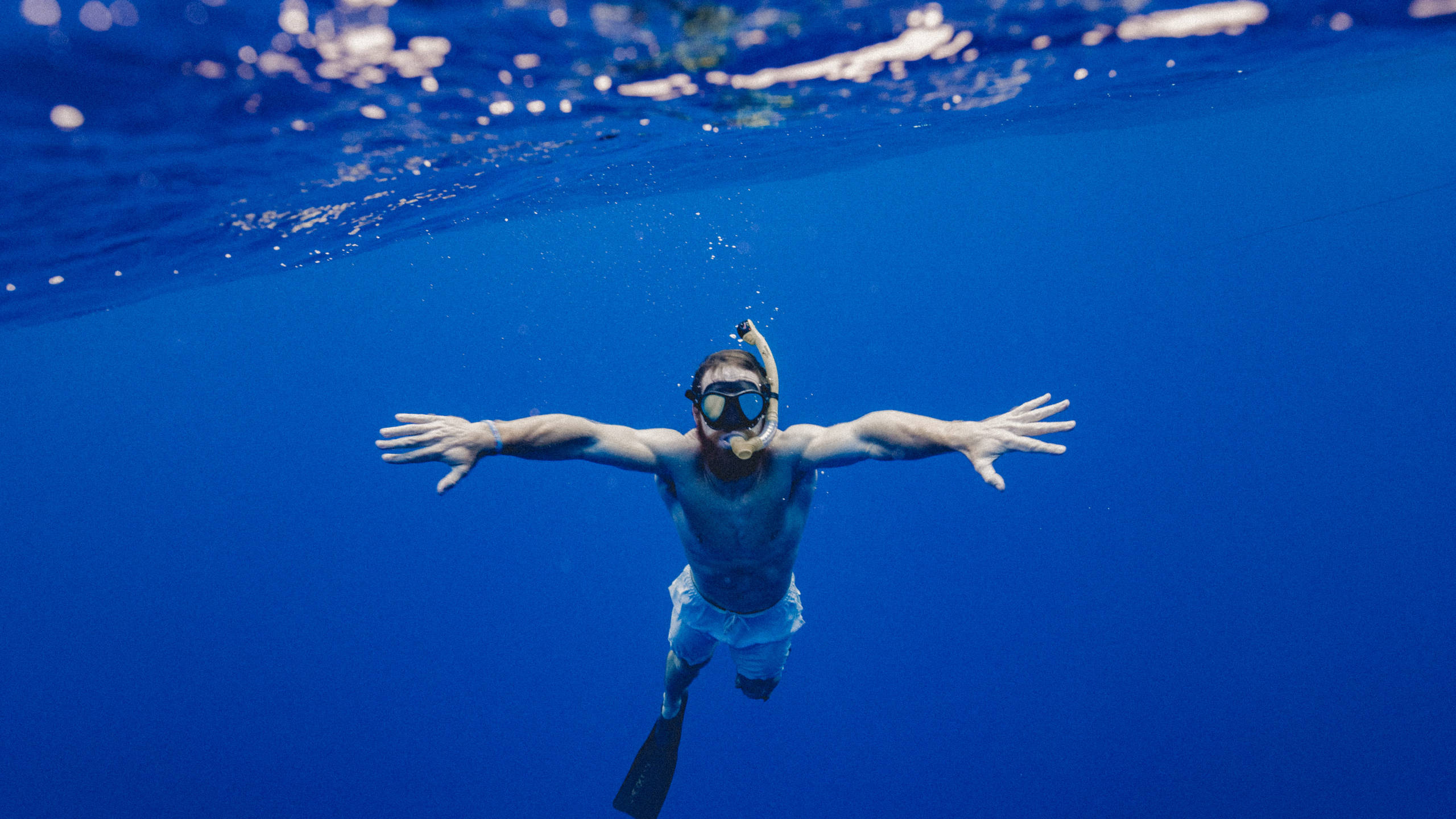 Swimming 桌布 壁紙 — 一名浮潛者在陽光照射的水面下伸展雙臂，滑過清澈湛藍的海水。