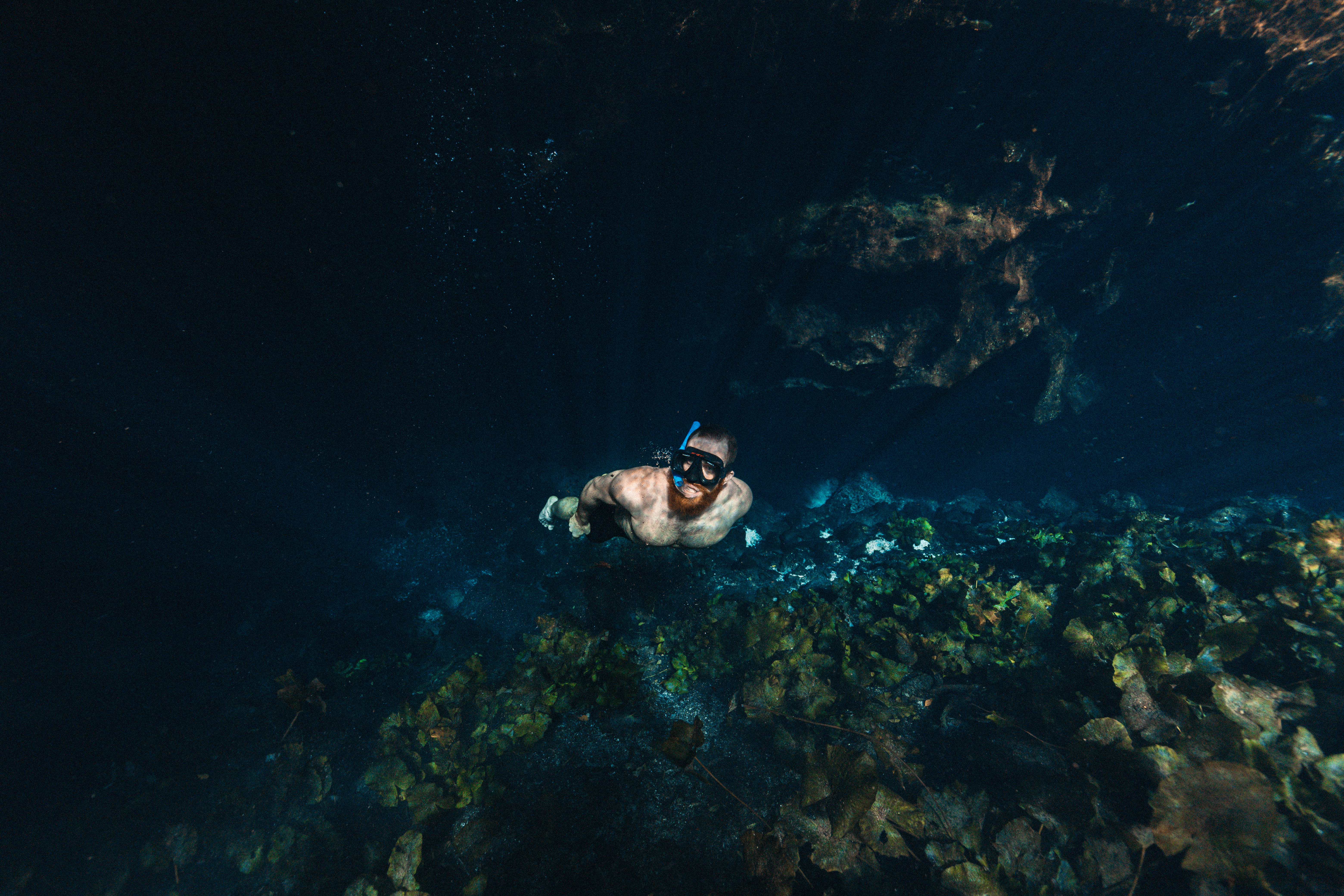 Snorkeling 桌布 壁紙 — 一名浮潛者在黑暗的水底洞穴中滑行，洞穴有岩石牆壁和散落的樹葉。