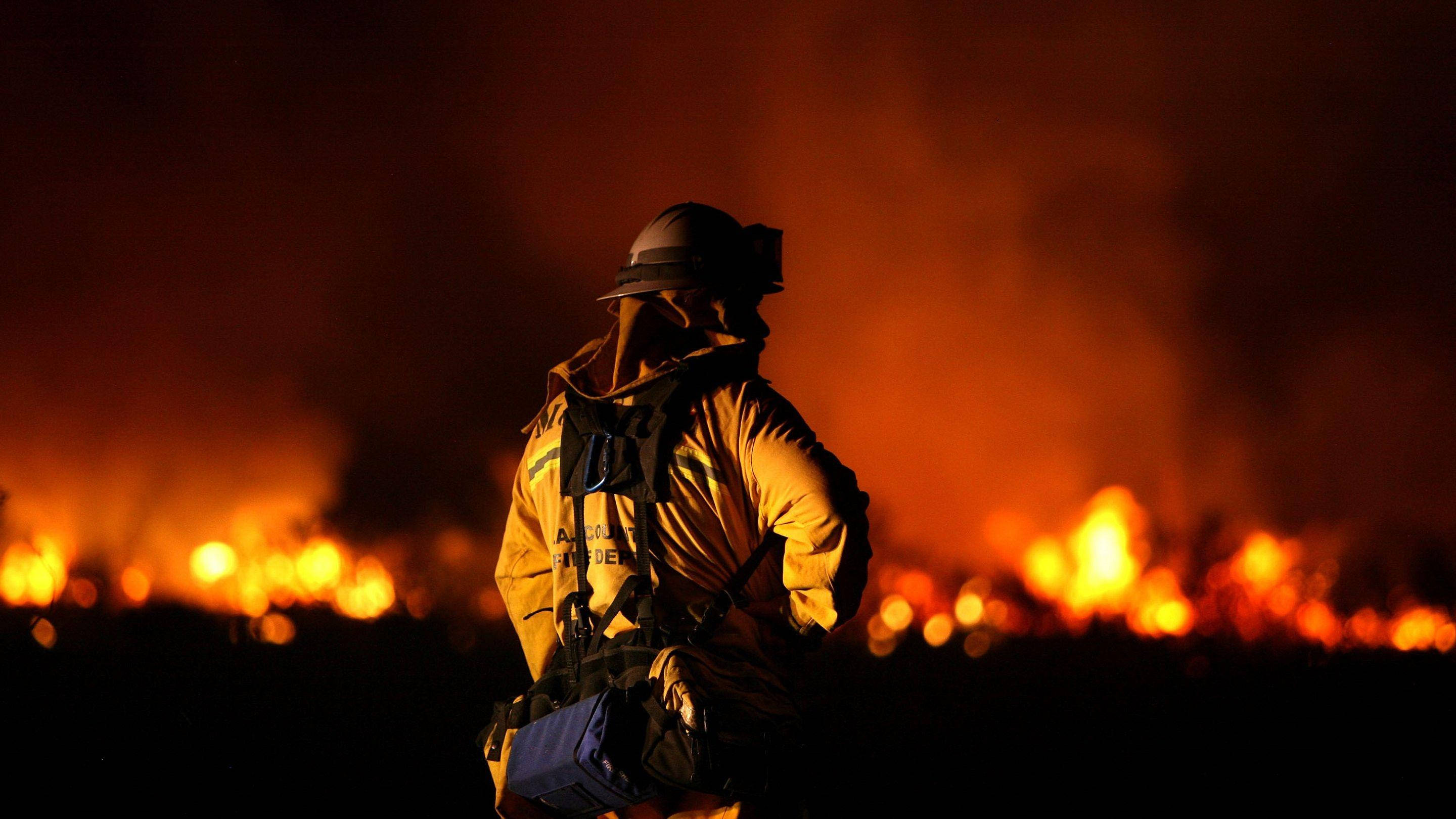 Firefighter 桌布 壁紙 — 一名消防員在夜間以剪影形式站立，背後是肆虐的野火、熾熱的火焰和濃煙。