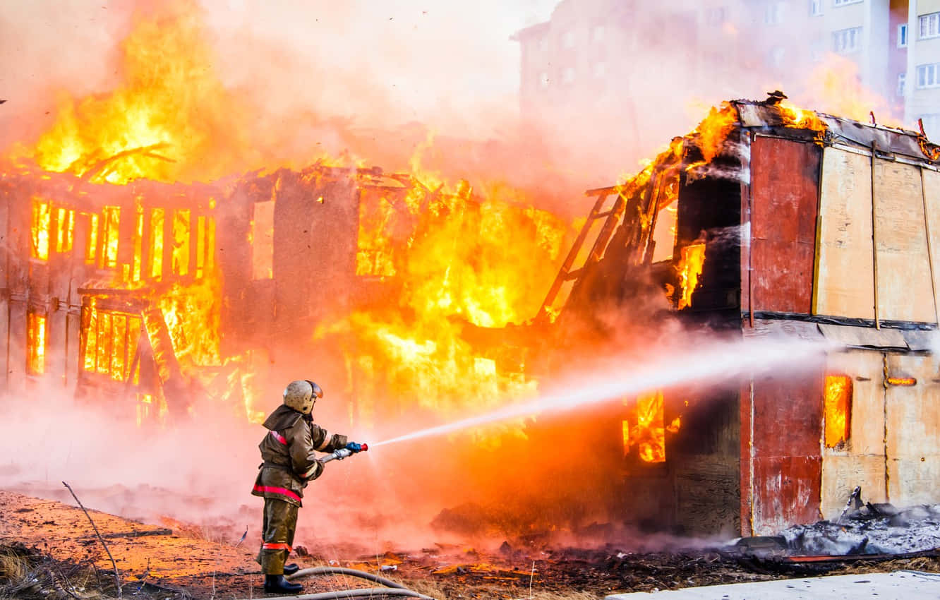 Firefighter 桌布 壁紙 — 一名消防員正在撲救一場猛烈的大火，大火正吞噬著一棟燃燒中的建築物，伴隨著強烈的橙色火焰和濃煙。