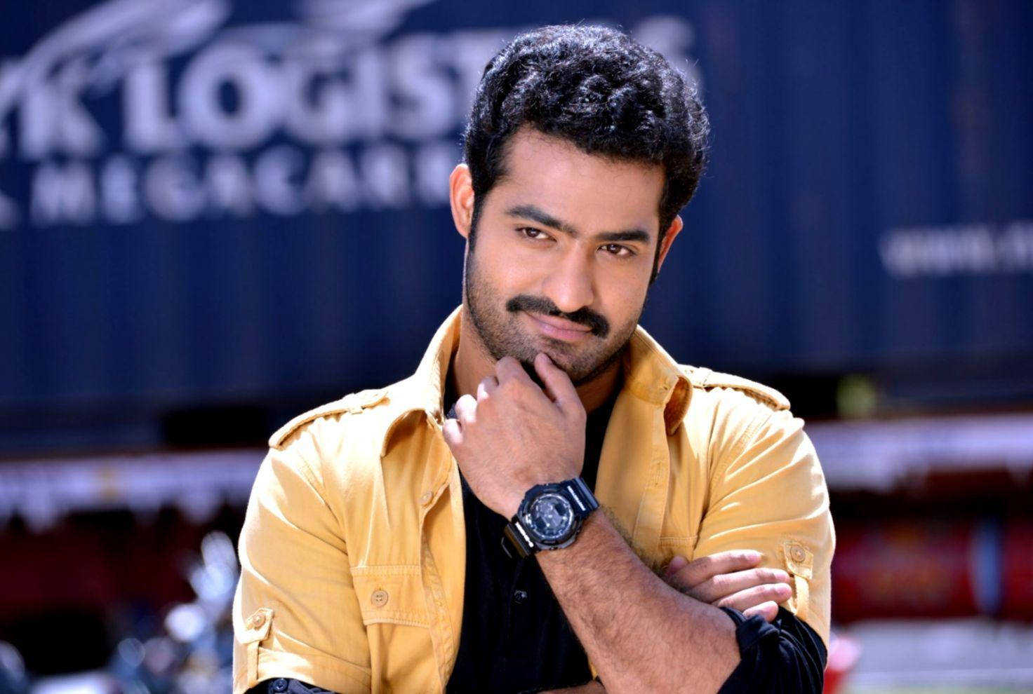 jr ntr 桌布 壁紙 — 一名深色捲髮、留鬍子的男士，身穿黃色襯衫和黑色手錶，手托下巴若有所思地擺姿勢。